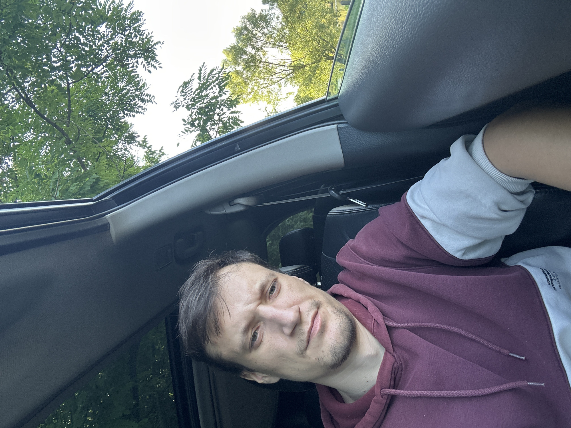 A man is sitting in the passenger seat of a car, looking directly at the camera. The car appears to be parked, possibly on a street in Makiv, Ukraine, as the lush green trees and bright sky visible through the car windows suggest an outdoor, possibly rural or suburban setting. The man is wearing a maroon and white color-block hoodie. His hair is dark brown and styled casually. The interior of the car is visible, including the car door and a portion of the dashboard. The image is taken from a selfie perspective, with the man's face and upper body filling most of the frame.
