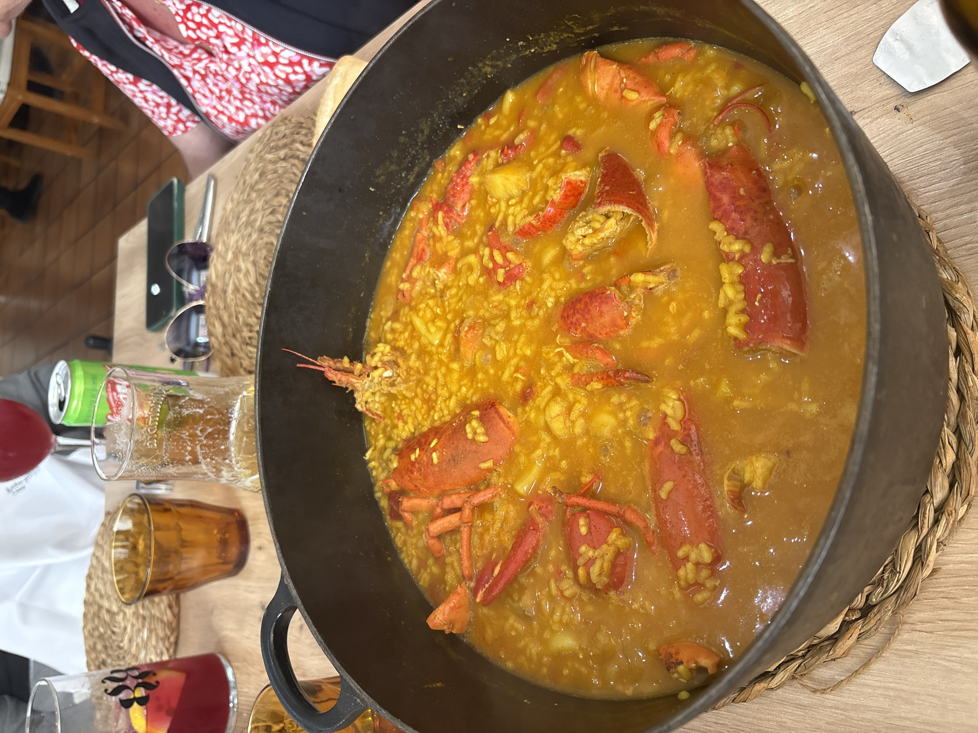 A high-angle shot depicts a paella, primarily featuring lobster, in a dark, possibly cast-iron, pan or paellera. The paella rests on a woven placemat on a wooden table. Several glasses and a can are visible on the left side of the frame. A person wearing a red and white patterned shirt is partially visible in the top left corner. A pair of sunglasses and a smartphone are also present on the table. The setting appears to be indoors, likely a restaurant. Based on the description provided, the location is Córdoba, Spain, given the mention of paella, suggesting a Spanish restaurant.