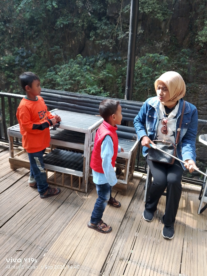 An outdoor scene in Sukadana, Indonesia, shows three individuals on a bamboo plank deck with dense green foliage and rocky terrain in the background.
On the right, an adult female is seated, wearing a light brown hijab, a blue denim jacket over a white patterned shirt, dark pants, and dark athletic shoes. Sunglasses are hooked onto her collar, and a dark, textured handbag rests on her lap. She holds a white extendable rod and is looking towards the two boys in front of her with a focused expression.
In the center, a smaller boy stands, facing the seated woman. He wears a red puffer vest over a light blue long-sleeved shirt, blue jeans, and dark sandals. To his left, a taller boy stands, oriented to the left and looking away from the camera and the other two individuals. He is dressed in an orange long-sleeved shirt with a graphic print, blue jeans with white text on the left leg, and dark sandals, holding a small dark object in his hands.
Two rustic, multi-tiered wooden structures with corrugated metal tops are positioned between the children and the woman. A dark metal railing is visible along the left edge of the bamboo deck, and a dark vertical pole extends upwards from the background foliage on the right.