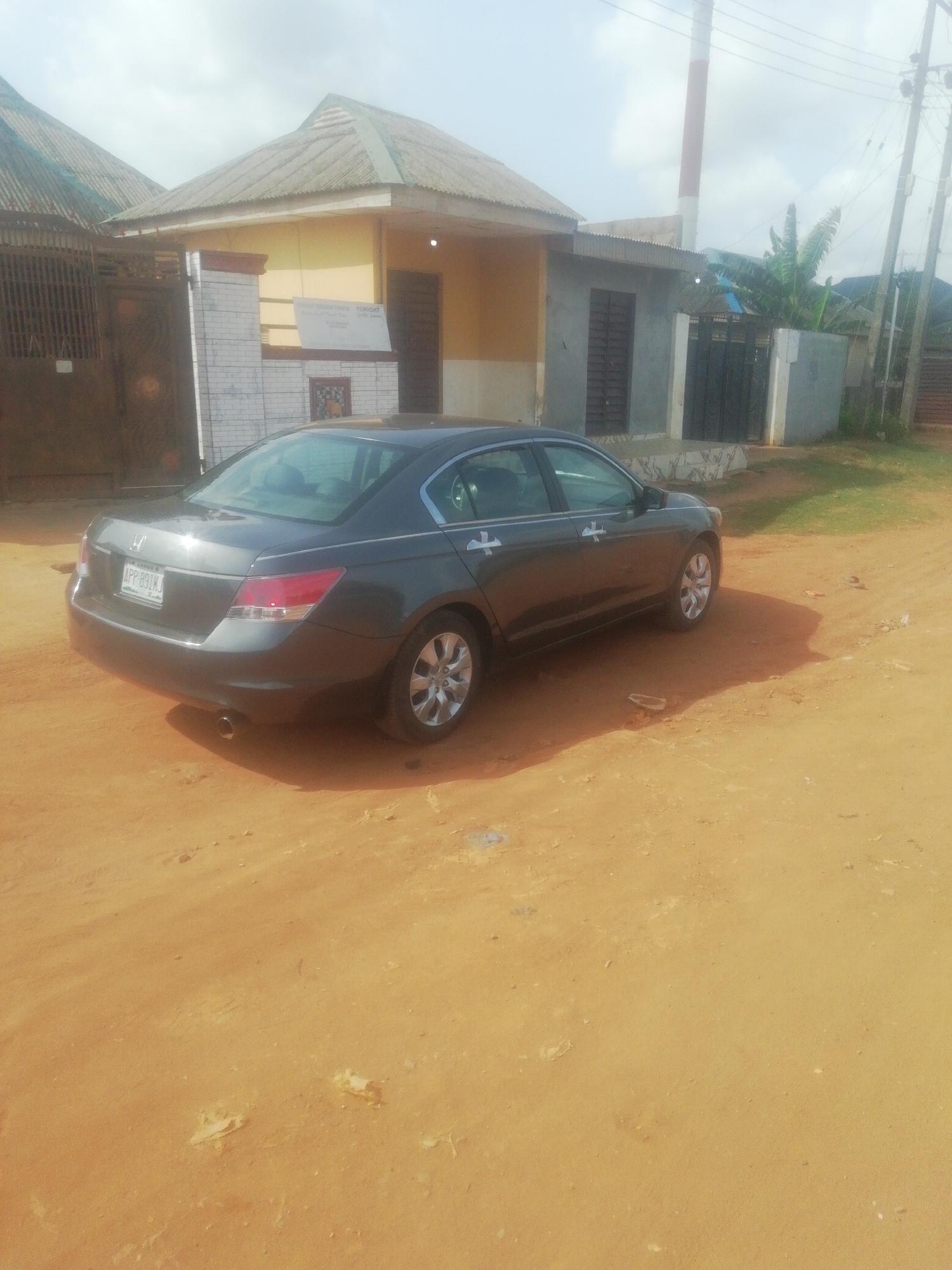 The image depicts a gray Honda Accord parked on a dirt road in a residential area. The location appears to be in Unknown, Unknown. The car is facing away from the camera, with its rear visible. Two residential buildings are visible behind the car, one with a yellow facade and the other with a gray facade. A utility pole with power lines is also visible. The sky is overcast.