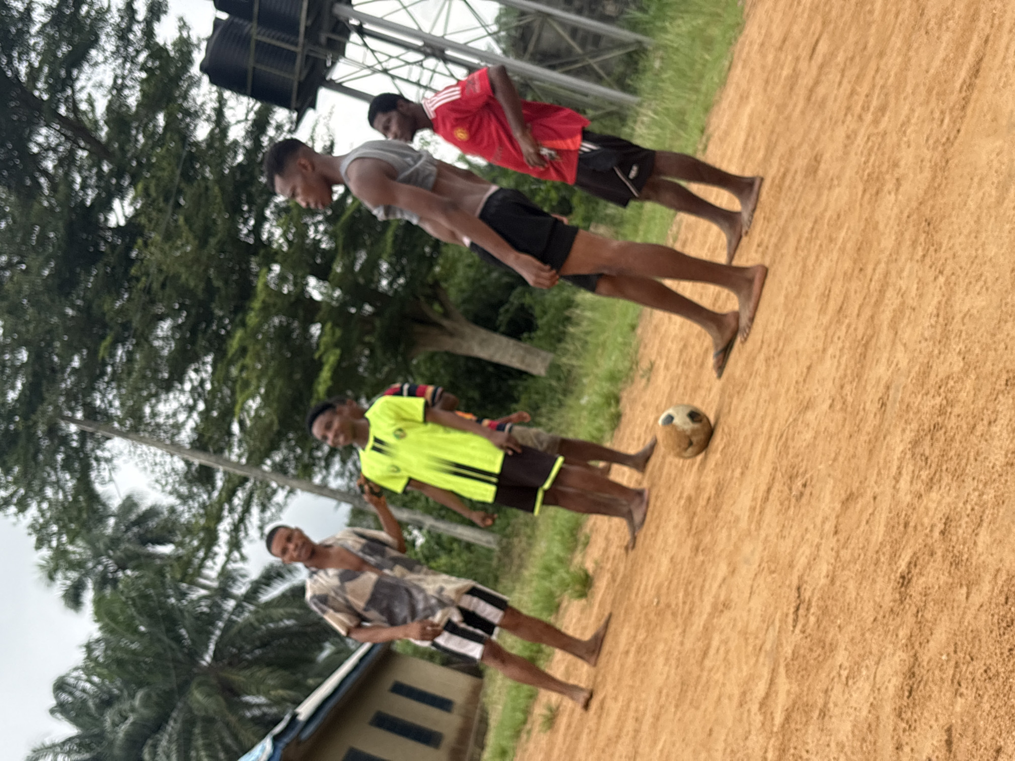 The image shows four young men on a dirt field in Ikot Ekpene, Nigeria. From closest to furthest, the men wear: a patterned short-sleeved shirt and black and white shorts; a yellow shirt with black trim and brown shorts; a grey tank top and black shorts; and a red Manchester United jersey and black shorts. They are all barefoot. A soccer ball sits on the ground in front of the second man. A building and trees are visible in the background. An overhead metal structure is present behind the first man. The field is red dirt. The sky is overcast.