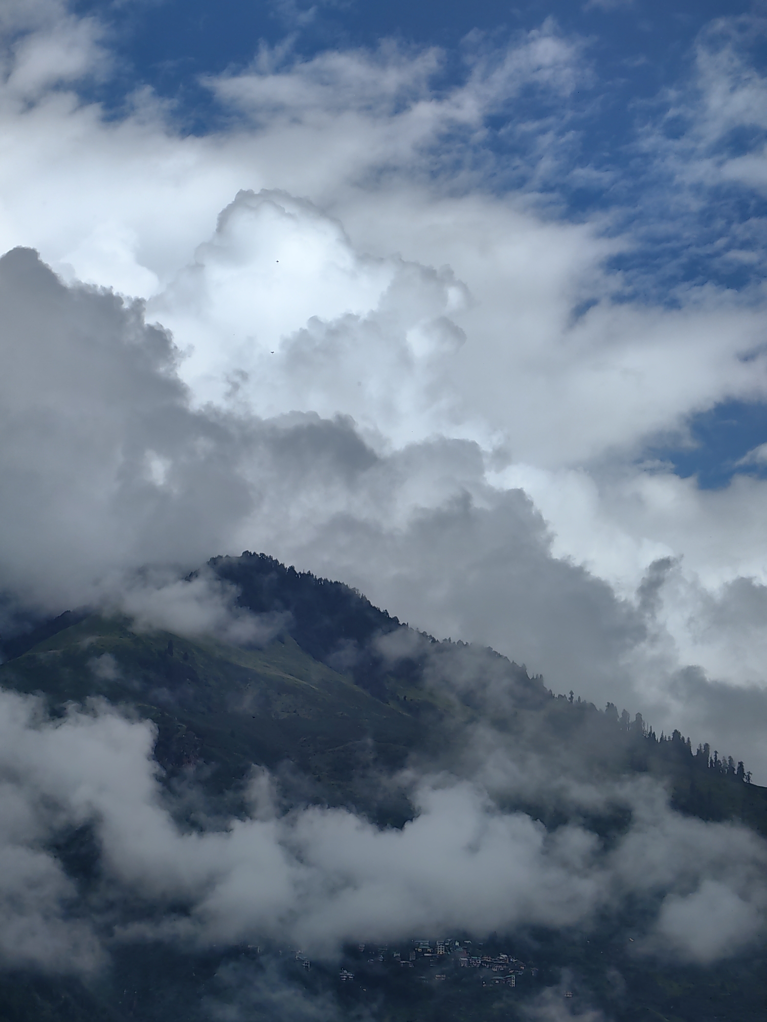 The image depicts a mountainous landscape, likely in Vashisht, India, characterized by dramatic cloud formations and lush green slopes. The sky is a dynamic mix of deep blue and thick, white and gray clouds, with some smaller clouds scattered throughout. The upper portion of the image is dominated by these clouds, some appearing cumulus and others more wispy.
Partially obscuring a steep, forested mountainside are dense layers of clouds, creating a sense of depth and atmosphere. The upper edge of the mountain is lined with a dense growth of dark green coniferous trees, appearing as a sharp silhouette against the lighter sky and clouds. Below the treeline, the mountain slopes are covered in vibrant green grass, with patches of darker shadow indicating rugged terrain.
Towards the bottom of the image, nestled on the lower slopes of the mountain, a small cluster of buildings is visible. These appear to be a village or settlement, with structures built close together. The buildings are a muted palette of browns and grays, blending with the natural environment. The presence of these structures suggests human habitation within this natural setting. The overall scene is one of a powerful natural environment with a subtle human presence, shrouded in a dramatic sky.
