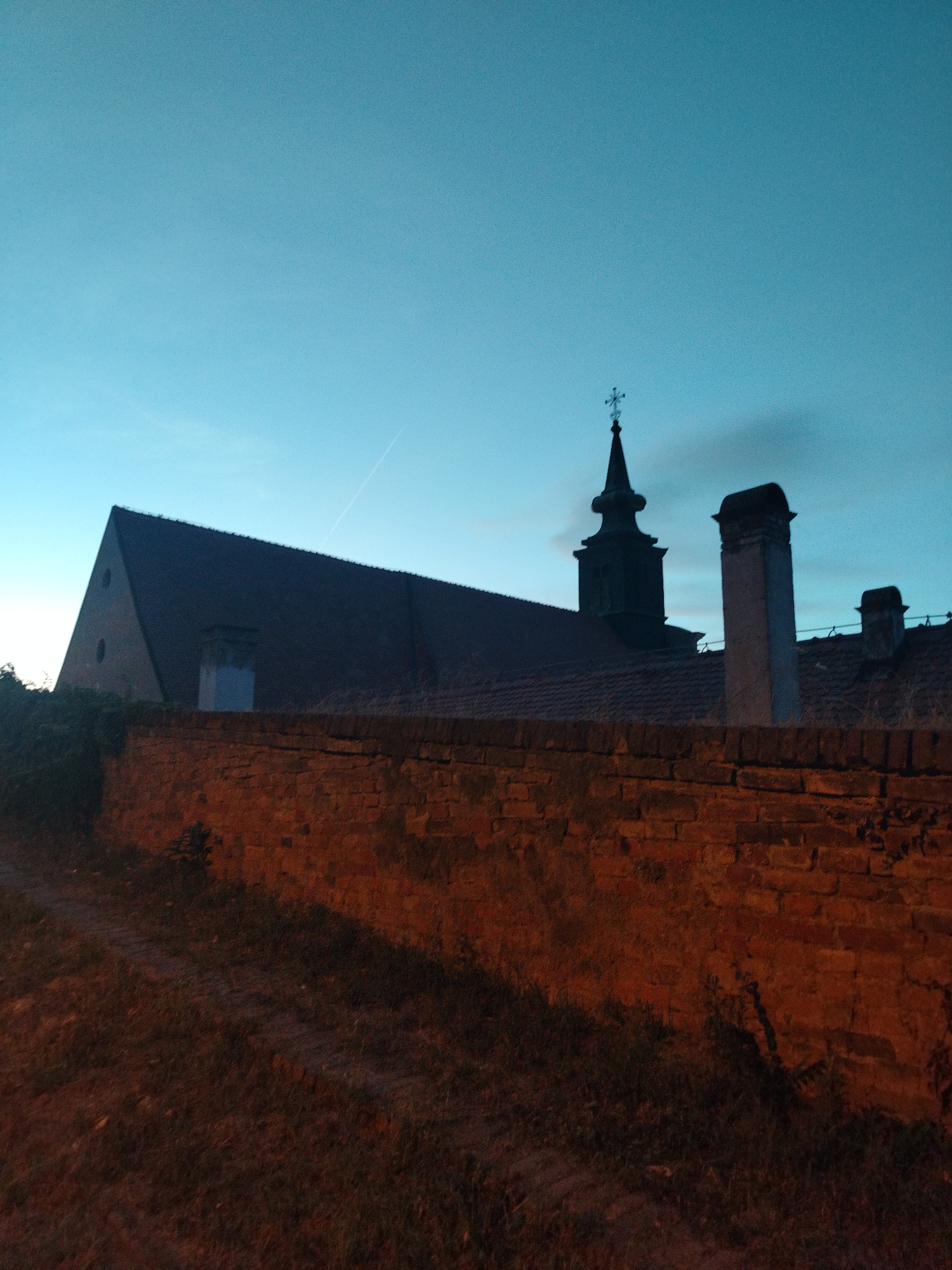The image shows a brick wall in the foreground, with a building and a church steeple visible behind it. The sky is a deep blue, indicating either dawn or dusk. The building appears to be old, with a red tile roof and a triangular gable. There is a church steeple with a cross on top, suggesting it is a place of worship. The brick wall is weathered and shows signs of age. The ground in front of the wall is covered in grass and dirt. A faint contrail is visible in the sky. The lighting suggests a low light condition, possibly early morning or late evening. The scene is devoid of people, with no discernible actions or interactions taking place. The overall impression is one of quiet and stillness. This scene is consistent with the older parts of Petrovaradin, Serbia, which is known for its historic architecture and fort.