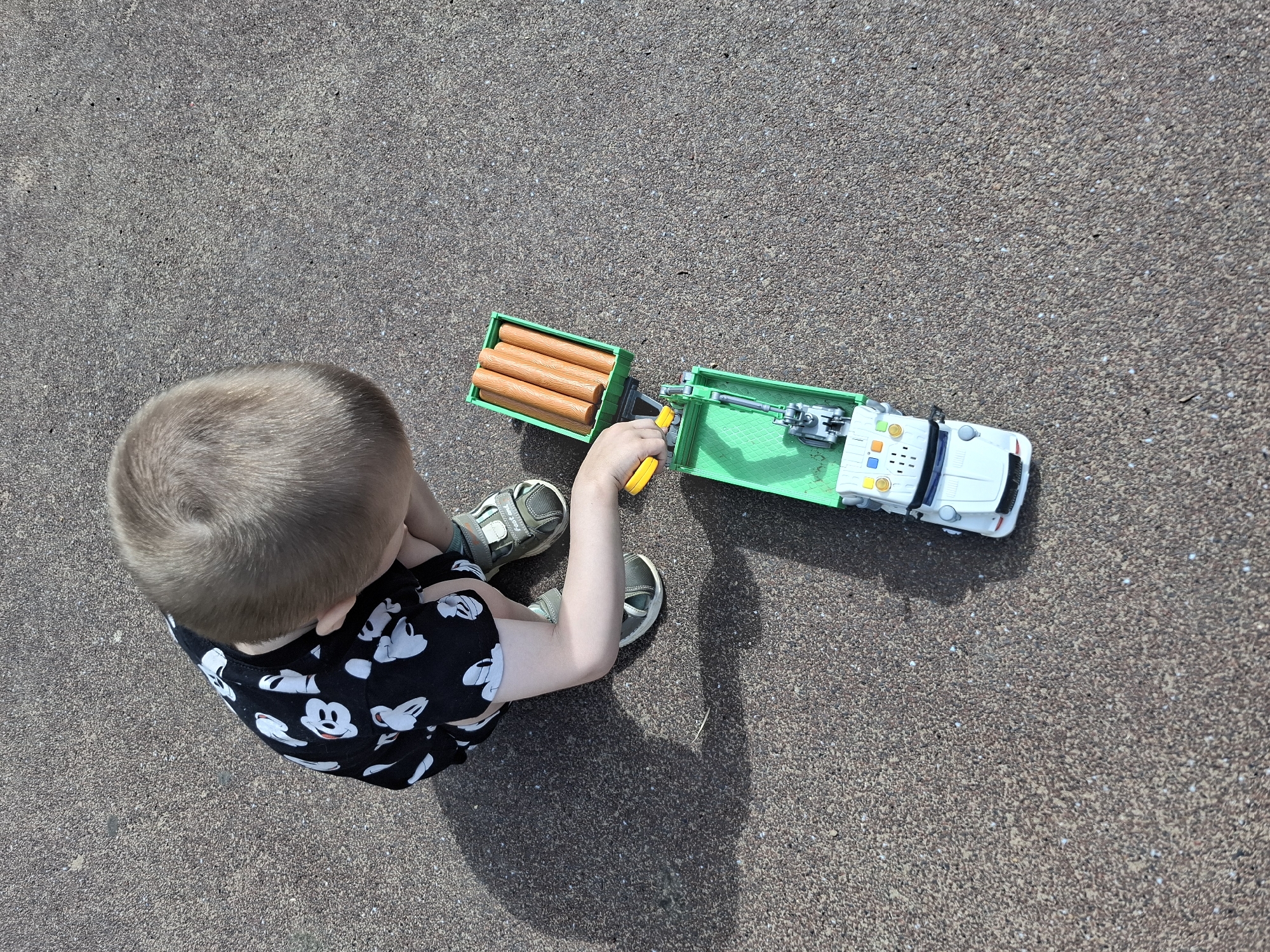 An overhead shot shows a young child with blonde hair, wearing a black t-shirt with white Mickey Mouse figures and grey sandals, kneeling on a paved surface that appears to be asphalt or a similar composite material, possibly in a park or on a sidewalk in Chaikovsky, Russia. The child is playing with two green toy trucks. One truck, a white cab with a green bed, has a grey mechanical arm. The other truck has a green bed filled with five brown, cylindrical objects, possibly representing logs or firewood. The child's left hand is reaching for the mechanical arm of the first truck, holding a yellow lever or control. Sunlight casts a distinct shadow of the child and toys on the ground to the left. The ground surface is textured with small, dark and light colored aggregate.