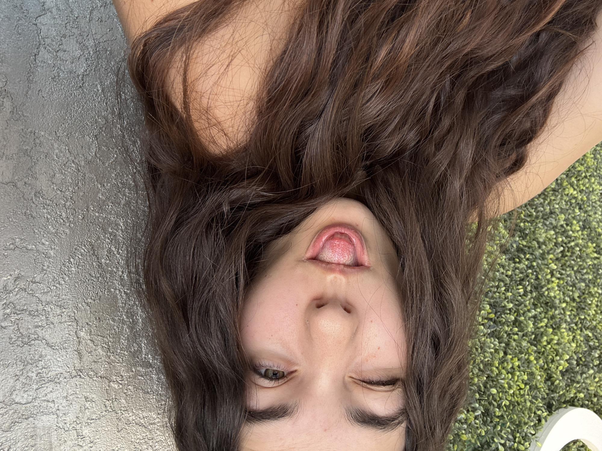 A young woman with long, wavy brown hair is upside down, sticking out her tongue, and winking. She is lying on a grey concrete surface on the left side of the frame and a green, leafy surface on the right side of the frame. Part of a white object is visible in the lower right corner. The exact location in Plovdiv, Bulgaria, cannot be determined from the image.