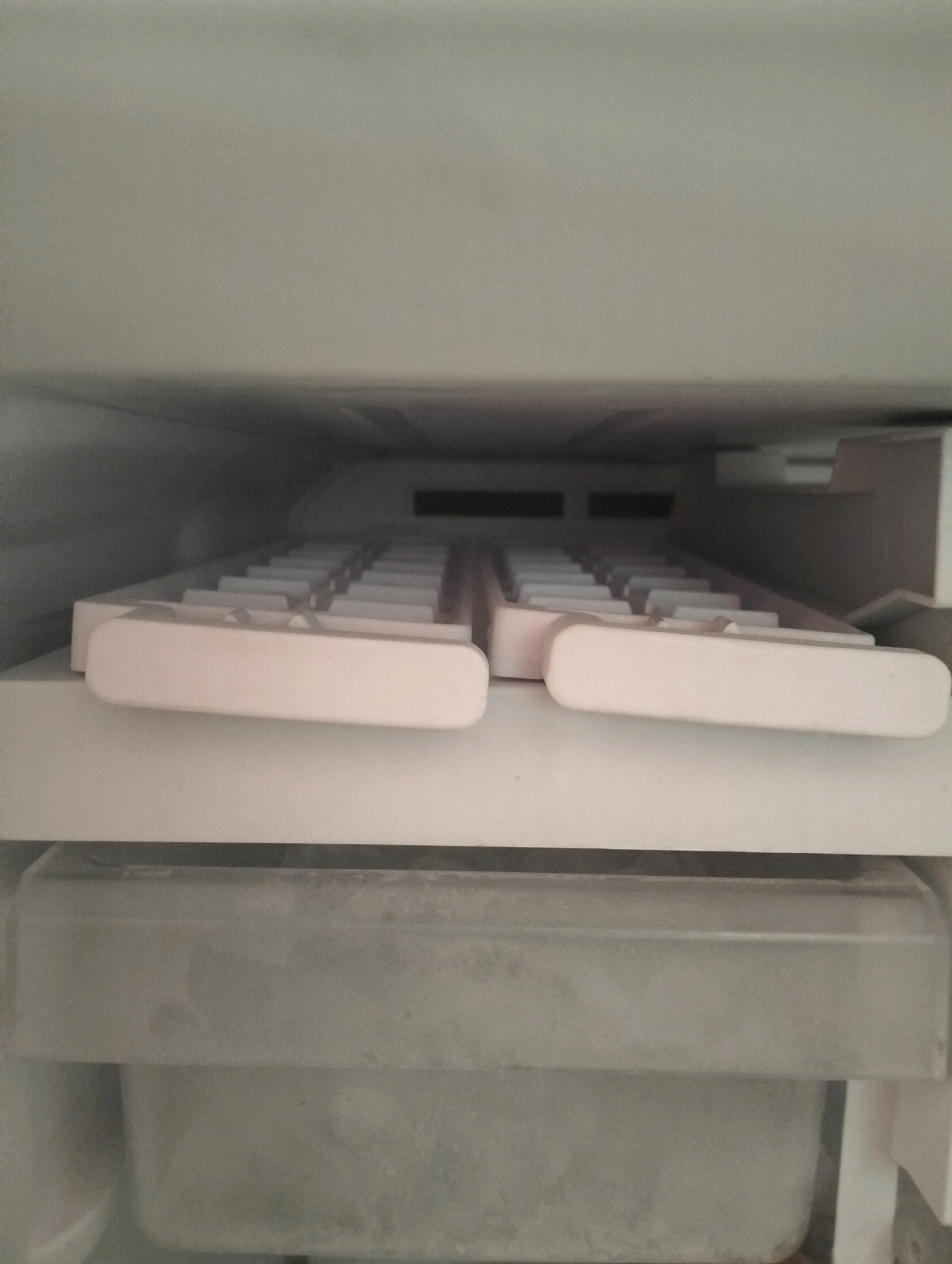 An interior view of a freezer compartment shows a white plastic ice cube tray with multiple rectangular molds, some of which appear to be filled with ice. The tray is positioned on a white ledge, with a grey plastic bin visible beneath it. The freezer walls are white and appear slightly frosted. The location is Tarai Bangun, Indonesia.