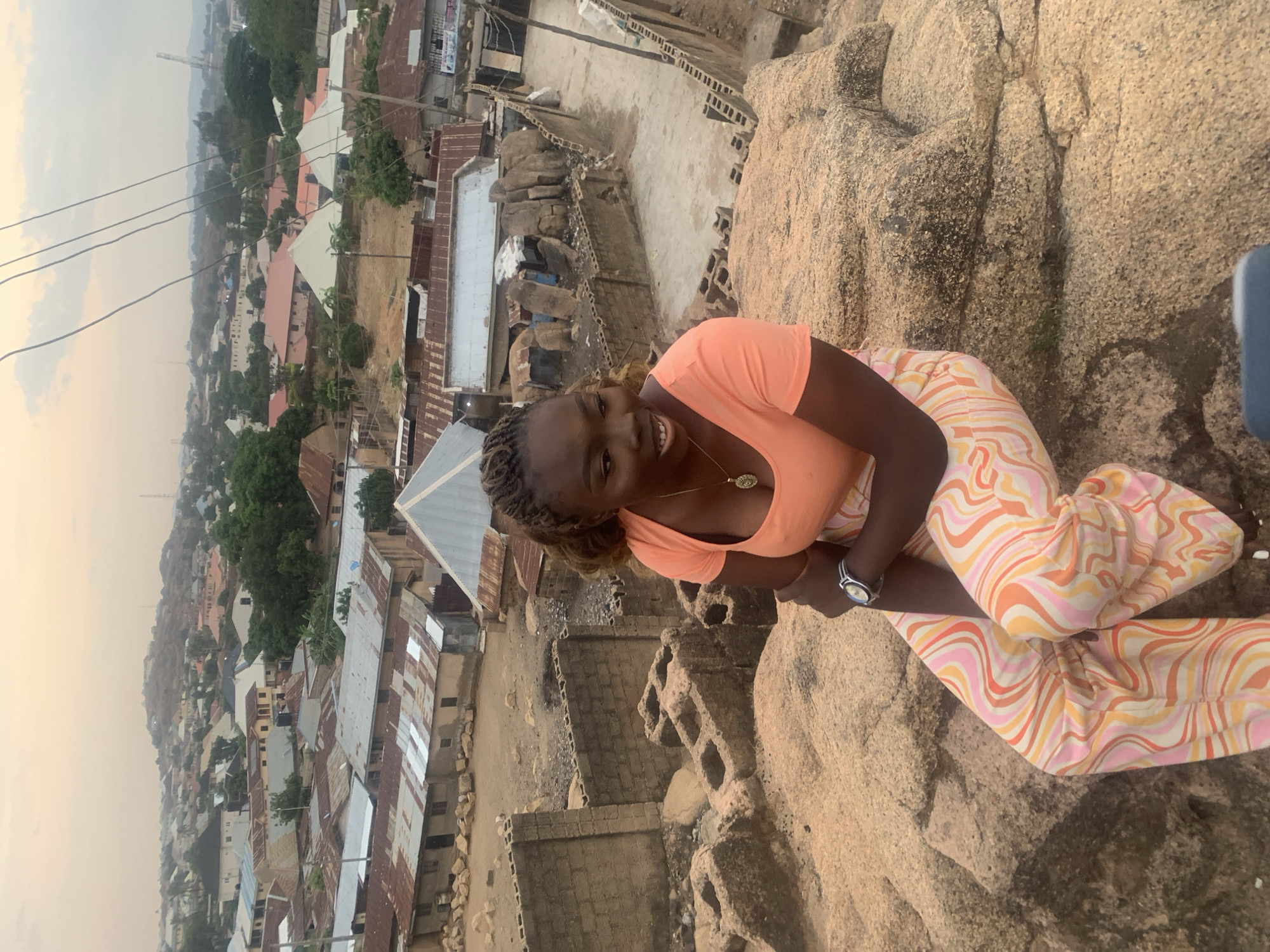The image shows a woman sitting on a large rock formation, smiling, in the city of Jos, Nigeria. She is wearing an orange shirt and patterned pants. In the background is a dense urban landscape with buildings of varying heights and designs, many with corrugated metal roofs. The sky is overcast with a hazy appearance. The rock formation she is sitting on has a rough, textured surface.