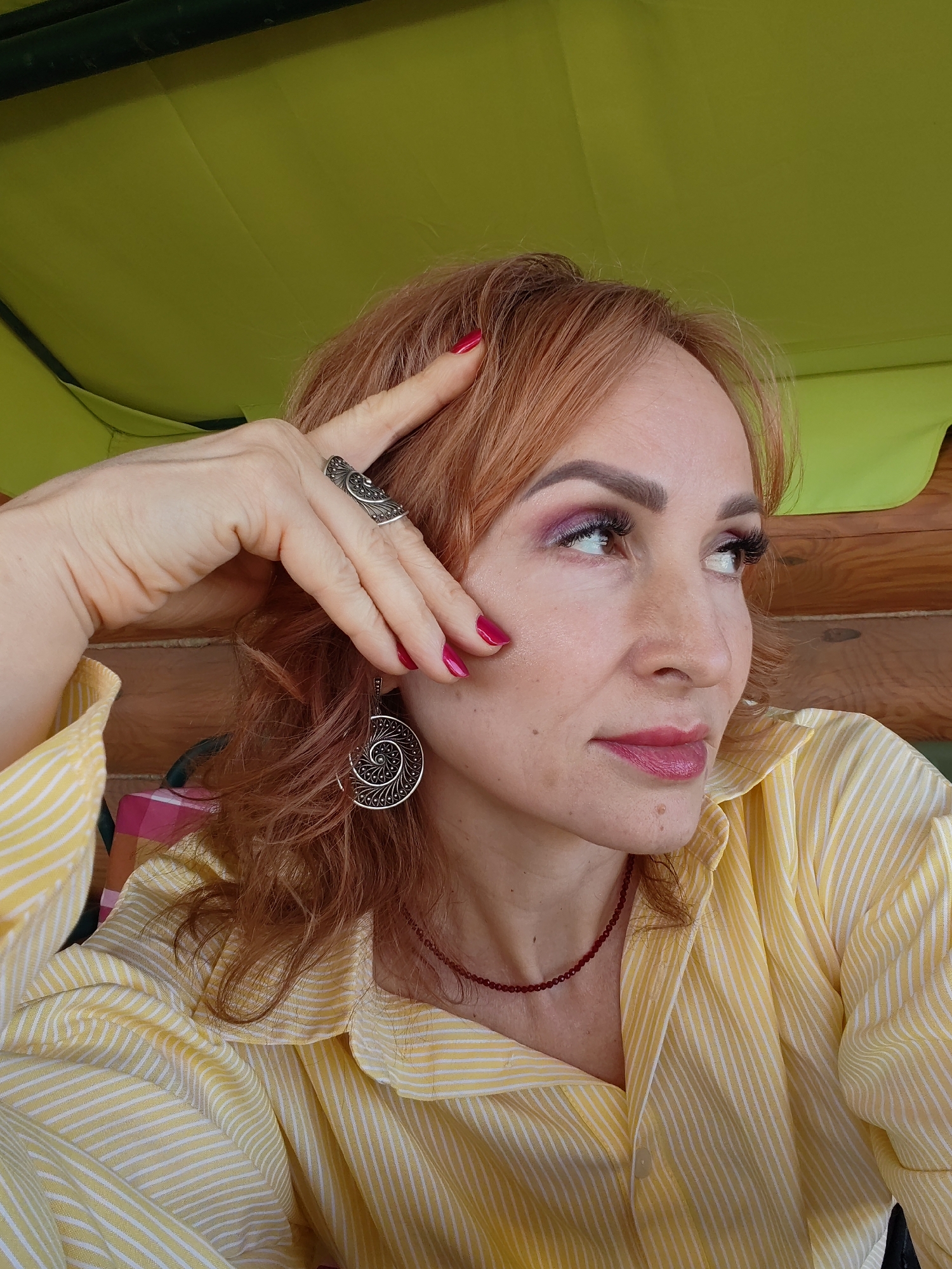 A woman with reddish-blonde hair is pictured in a close-up shot. She is wearing a yellow striped shirt and is adorned with a large silver ring and matching earrings. Her nails are painted a bright pink. She is looking to the side with a contemplative expression. She is wearing pink eye shadow and lipstick. The background includes green and wood tones. The location is Старое Михайловское, Russia.