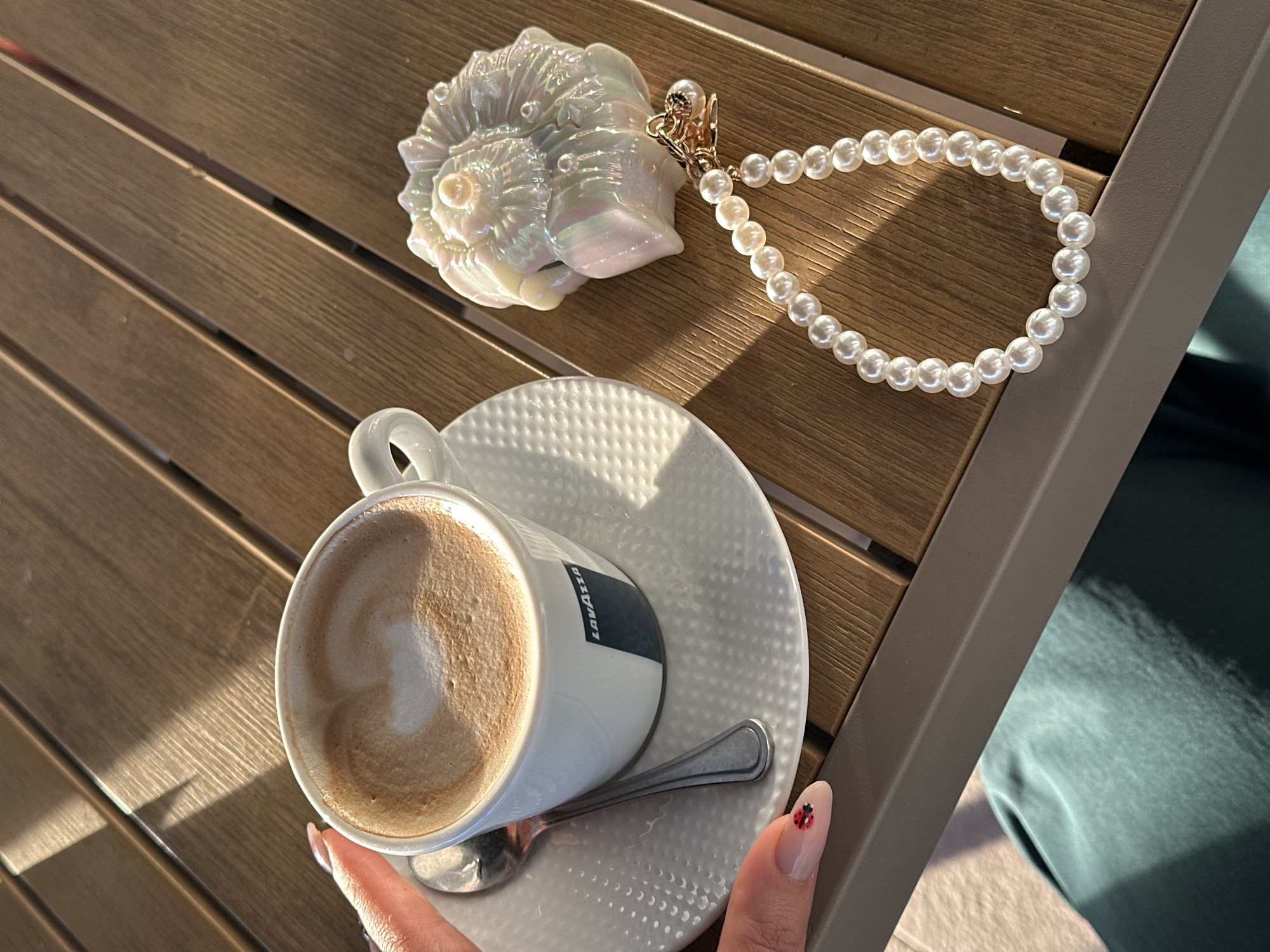 The image captures a close-up, high-angle view of a person's hand holding a coffee cup on a wooden slatted table in bright sunlight. The setting appears to be an outdoor cafe or patio.
The primary subject is a white ceramic coffee cup, branded "LAVAZZA" in blue text, filled with a latte or cappuccino featuring a heart-shaped latte art design. It rests on a white textured saucer, with a silver spoon placed beside it. A person's hand, with manicured nails including a small red ladybug design on the thumb, holds the cup.
On the table, to the upper right of the coffee, lies a decorative, iridescent white seashell ornament. Attached to the seashell is a string of white pearls with a gold clasp. The wooden table has parallel slats, and strong sunlight casts distinct shadows, indicating a bright daytime environment. A grey metal frame or leg of the table is visible on the right, and hints of green fabric (possibly clothing) and light-colored flooring are seen in the background.
The scene suggests a relaxed moment, possibly a coffee break in a sunny location, consistent with a cafe in a coastal town like Bar, Montenegro.