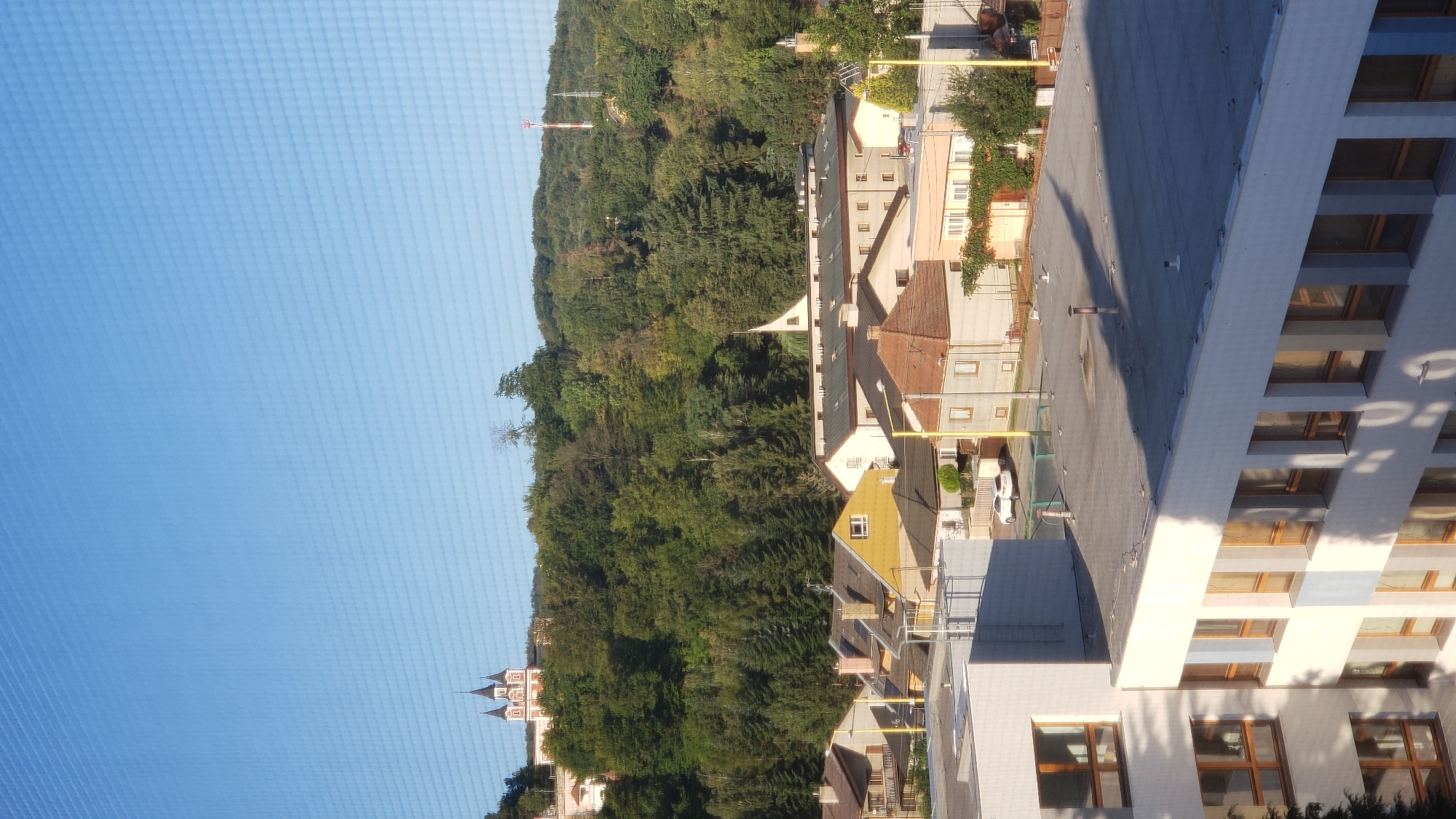 A sunlit, wide-angle photograph taken from an elevated perspective shows a residential area in Prešov, Slovakia, with a forest-covered hill and a church in the background. In the foreground, the top of a white building with multiple rows of windows is visible. The middle ground features a collection of houses with red-tiled roofs and yellow and beige facades. Beyond the houses, a dense forest covers a hill, with a distinct church structure featuring multiple spires visible at the crest. A radio tower is also visible on the far right side of the hill. The sky is clear and blue. There are no people or animals visible in the image. The scene depicts a peaceful, suburban environment during daylight hours.