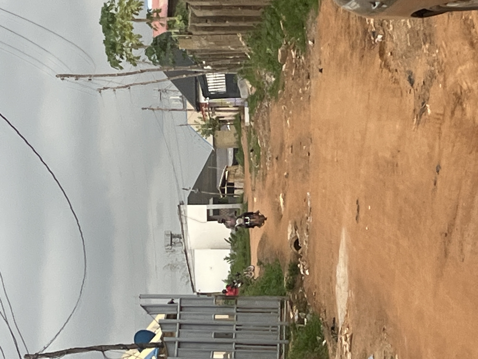 The image captures a ground-level view of a dirt road in a residential or semi-urban area, likely in Abuja, Nigeria. The unpaved road, composed of reddish-brown earth, dominates the right side of the frame and appears to recede into the distance.
Along the left side of the road, a series of simple, mostly single-story buildings with white or light-colored walls and dark roofs are visible. These structures are intermingled with patches of green vegetation and a few trees. Utility poles, some made of wood, line the roadside, supporting a complex network of overhead electrical or communication wires that crisscross the sky.
In the midground, a person is seen riding a motorbike away from the viewer, traveling down the dirt road. Further beyond, near the buildings, a few other indistinct human figures can be observed, one notably wearing red attire. The immediate foreground on the left features a metal fence-like structure and a blue circular object, while the top right corner shows part of a vehicle.
The weather appears overcast, indicated by the uniformly gray sky and the lack of strong shadows, suggesting daytime without direct sunlight. The scene depicts everyday activity in a local community, characterized by modest architecture and basic infrastructure. No visible text is present in the image.