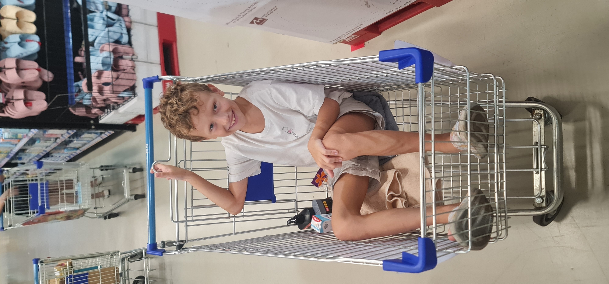 A young boy is sitting inside a metal shopping cart at a retail location in Đà Nẵng, Vietnam. The boy has fair skin and curly blonde hair, and he is wearing a white t-shirt and khaki shorts. He is smiling and looking at the camera, with one arm extended to hold onto the side of the cart. Inside the cart with the boy are various items, including a towel, a blue cushion, and personal care products. The surrounding environment is a well-lit store with aisles of merchandise on shelves. Other shopping carts are visible in the background.