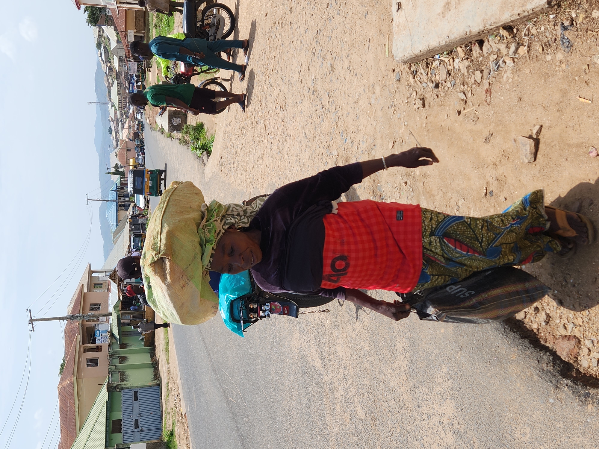 An older woman carries a large bundle on her head while riding a small moped on a paved road in a densely populated urban area. She wears a dark long-sleeved shirt, a red and black patterned garment around her waist, and a colorful patterned skirt. On her head is a large, bulky bundle wrapped in light green fabric. The moped is small and appears to be carrying goods or personal items as well. The road appears to be the main thoroughfare of a community or town, with buildings visible along both sides of the road. Other people and motorcycles are seen in the background. The overall setting suggests a bustling environment.