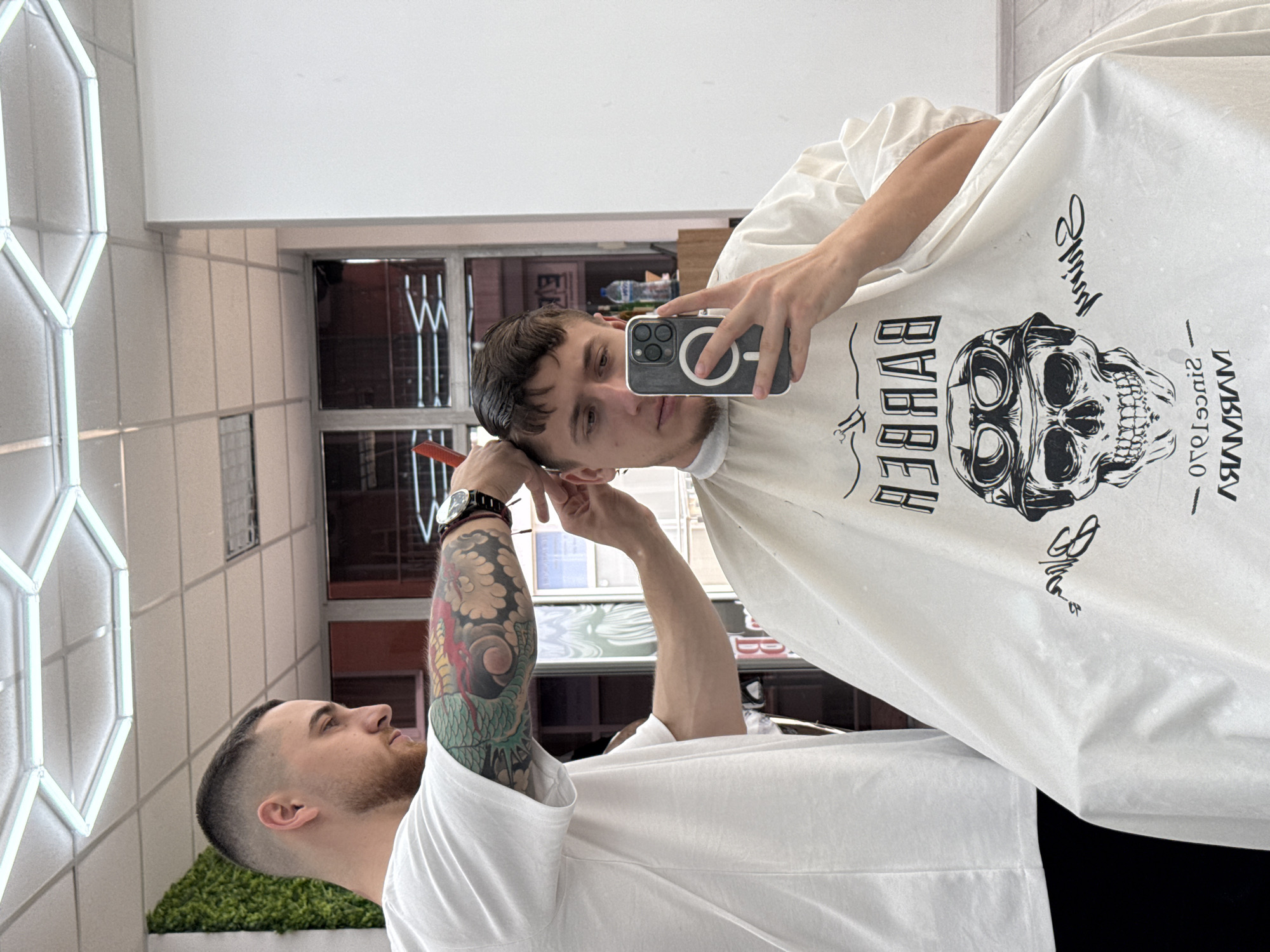 The image shows two men in what appears to be a barbershop. One man is seated, wearing a barber's cape with a skull graphic and the word "BARBER" printed on it, taking a selfie with his phone. The other man, a barber, is standing next to him, holding a comb and appearing to style his hair. The barber has a tattoo sleeve on his right arm and is wearing a watch. The shop's interior features tiled walls, a lighted geometric design, and a view through a window, suggesting a location in Sofia, Bulgaria. The windows show the reflection of a modern, urban environment with a building across the street.