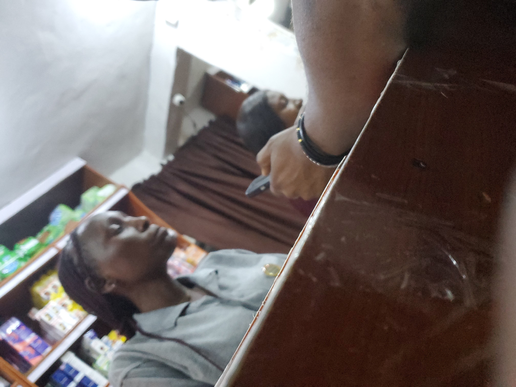 The image depicts an indoor scene, likely a small shop or convenience store, with shelves stocked with various products in the background. Two individuals are visible, both appearing to be female. One woman, in the foreground to the left, is wearing a light grey collared shirt and has her hair in braids. She is looking downwards and appears to be in profile, with the camera positioned slightly above her eye level. The other individual, partially obscured and further in the background to the right, is also looking downwards and appears to be holding a mobile phone. A hand wearing a black bracelet with gold accents is visible in the mid-ground, holding the phone. The environment suggests a retail setting, with a counter or display surface in the foreground. The overall lighting is somewhat dim, with a bright white area suggesting a light source or reflections on the ceiling or upper wall. The scene does not provide any specific landmarks or visual cues to pinpoint a precise location within Jos, Nigeria, beyond it being an urban commercial setting.
