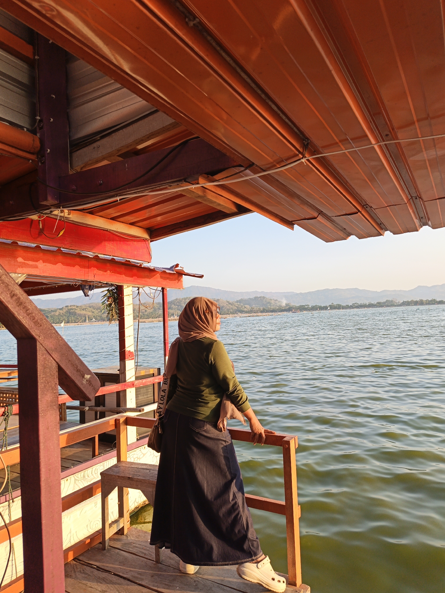 In Krakitan, Indonesia, a woman wearing a hijab and a long dark skirt stands on a wooden deck, looking out at a wide body of water. She is leaning on a wooden railing, her right hand resting on it. The water is calm, with small ripples on the surface. In the distance, a line of trees and hills forms the horizon, bathed in the warm light of what appears to be late afternoon or early morning sun. The deck is part of a structure with an orange roof supported by beams. A portion of a red railing and some wooden stairs are also visible on the left side of the frame. The woman carries a small brown bag over her shoulder. The scene is peaceful and scenic, with the natural landscape contrasting with the man-made structure.