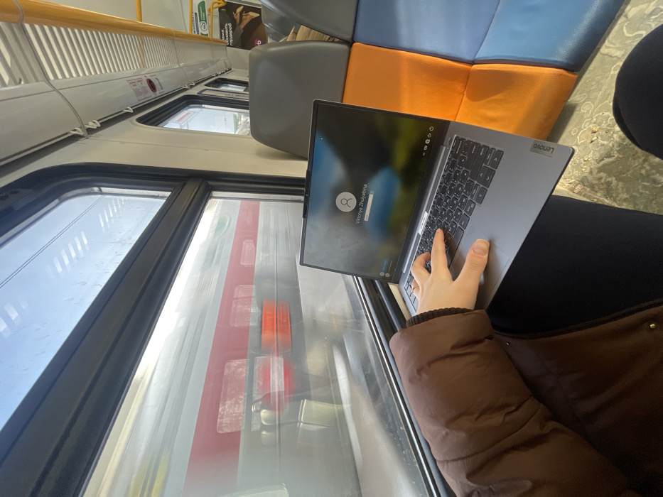The scene depicts the interior of a passenger train carriage, stated to be in the city of Бабеево, Russia. A person, observed from a first-person perspective, is seated, with their left arm in a brown puffed jacket and dark trousers visible. A silver-grey Lenovo laptop is open on the person's lap. The laptop screen displays a login interface featuring a generic profile icon, the name "Viktoriya Zhulyabina," and a password input field. The individual's left hand is positioned over the laptop's keyboard, poised for typing. The keyboard features both Latin and Cyrillic characters. Through the train window, the side of another passenger train is visible, characterized by a light grey body with a distinct horizontal red stripe; a motion blur suggests relative movement. The interior of the carriage includes windows with black frames, overhead panels, and visible padded seats in grey, orange, and blue upholstery. Sunlight illuminates the interior.