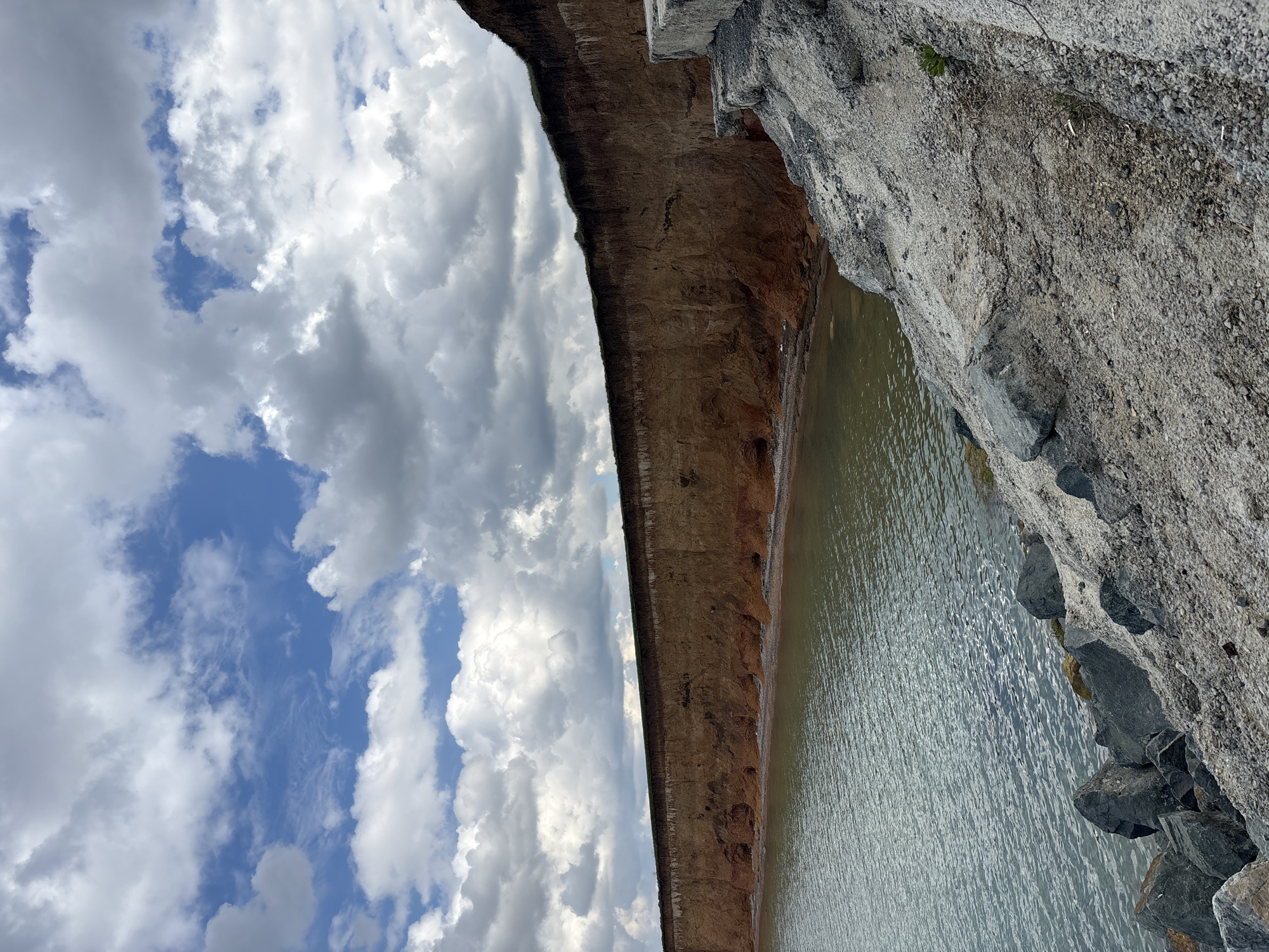 The photograph shows a body of water beside a cliff face under a cloudy sky. The cliff face has visible striations and appears to be composed of different layers of earth and rock. The body of water is calm with subtle ripples. The sky is filled with puffy white clouds, with patches of blue visible between them. The photograph appears to be taken at Николаевское сельское поселение, Ukraine, but no specific landmarks or identifying features are visible to confirm this. There are no people visible in the image.