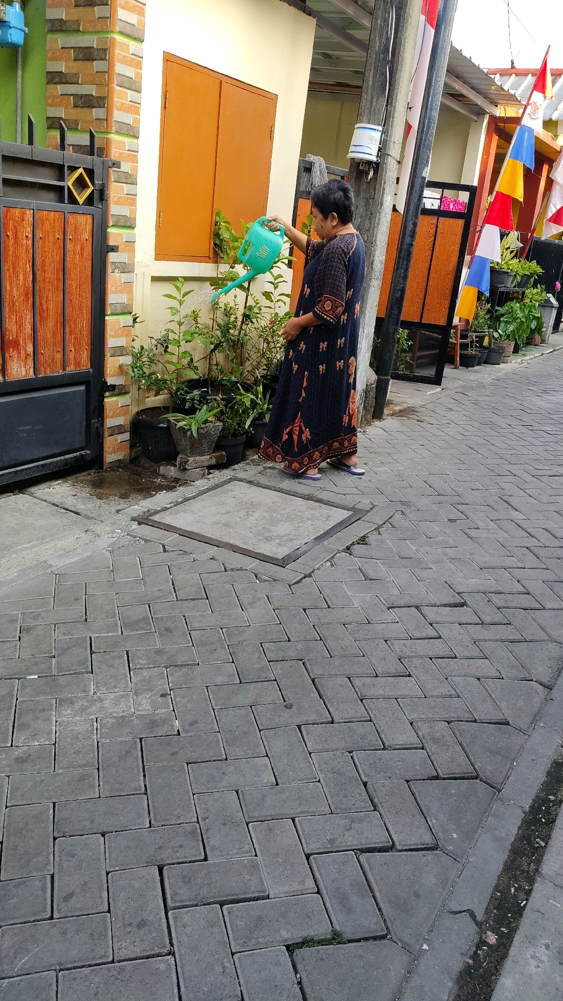A woman waters plants in front of a building in Tangerang, Indonesia. The woman is wearing a dark blue dress with a patterned design. She is holding a green watering can and watering a small bush. The building behind her is light yellow with orange window frames. In front of the building, there are several potted plants. The ground is paved with gray interlocking bricks. There is a black metal gate to the left of the woman, and a pole with flags to her right.