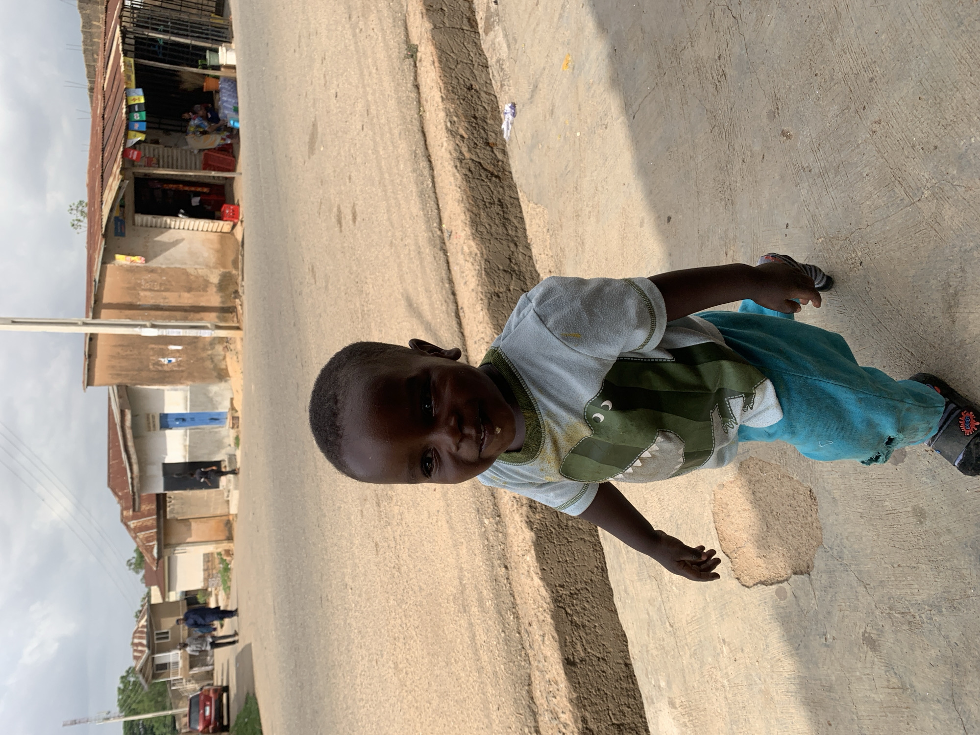 A young child stands in the street of Jos, Nigeria, looking directly at the camera. The child is wearing a short-sleeved shirt with a green crocodile on the front, light blue pants with a hole in the knee, and black sandals. The background includes a paved road with buildings lining the street. Some buildings are shops with goods displayed. People are walking on the street in the distance. The sky is overcast.