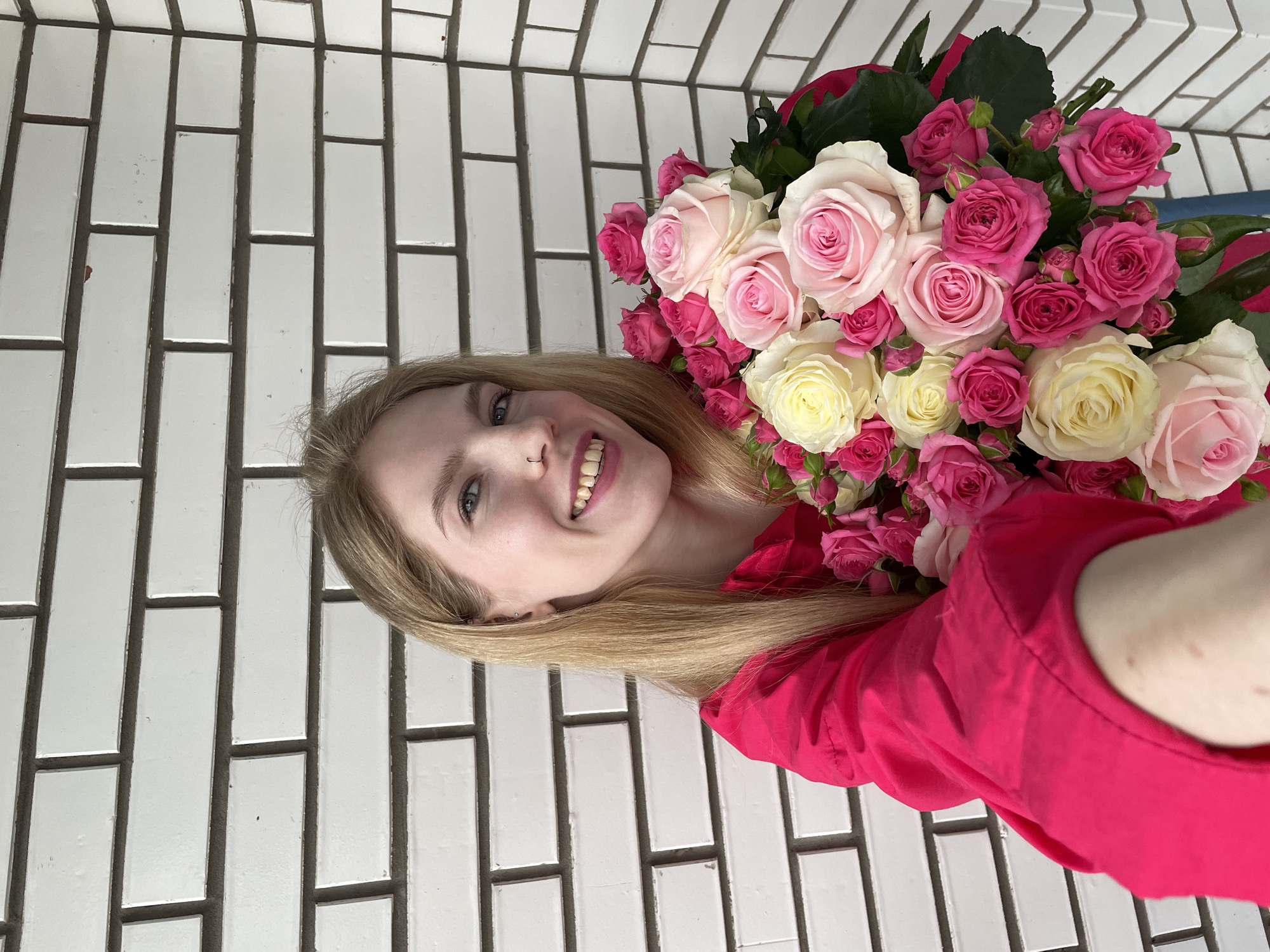 A young woman with blonde hair smiles at the camera while holding a bouquet of pink and white roses. She is wearing a bright pink top. The background consists of a wall tiled with white, rectangular tiles. There is no information to conclude this is Izhevsk, Russia.
