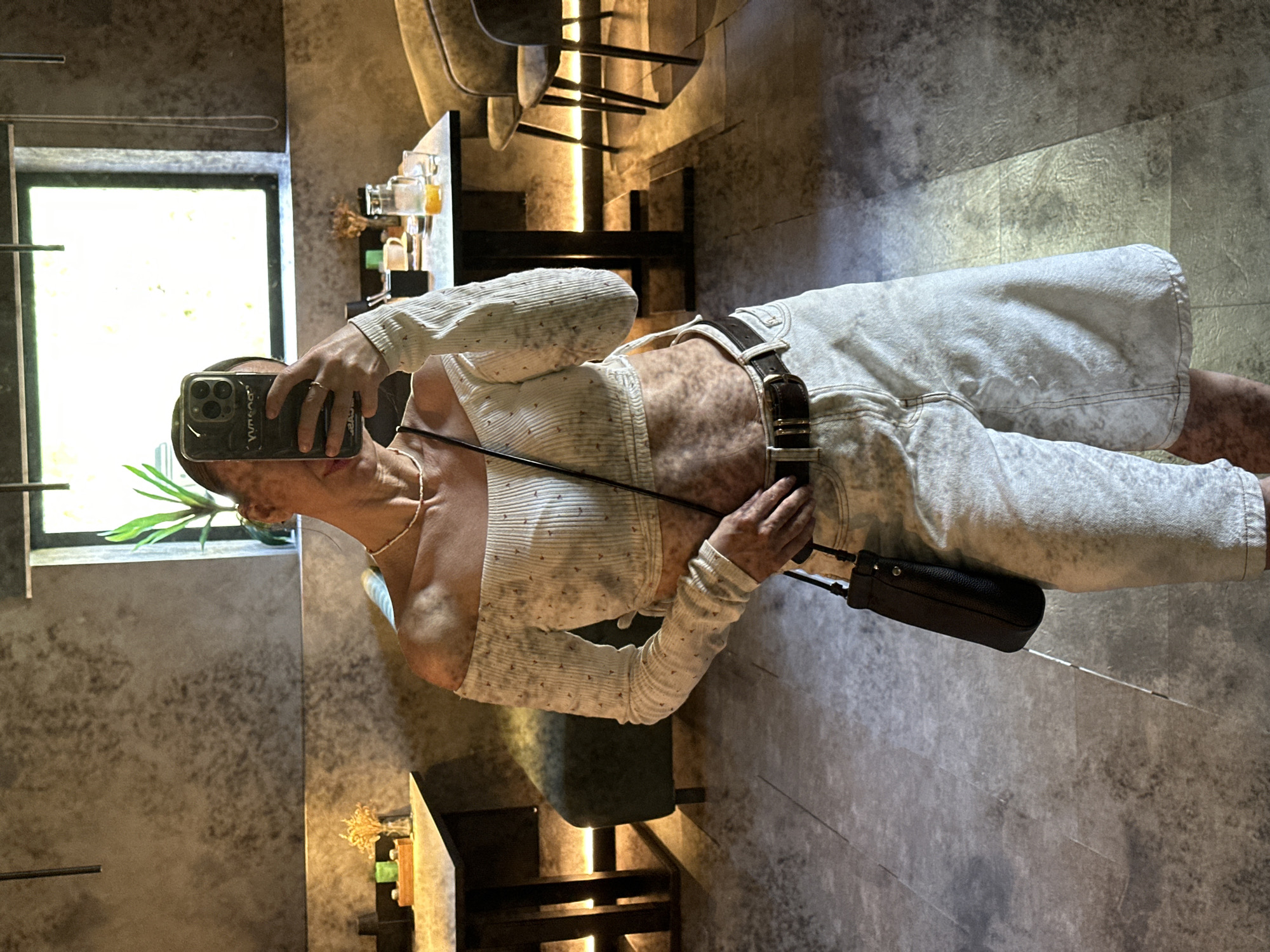 A person is taking a selfie in a room with a gray concrete-like wall. The person is wearing a white long-sleeved shirt, light-colored jeans with a brown belt, and a black purse. They are holding a smartphone in their left hand to take the picture. A window with greenery is visible in the background. The room has furniture, including chairs and tables. The location is Votkinsk, Russia.