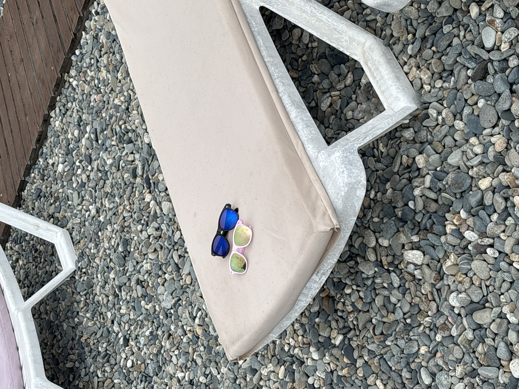 Two pairs of sunglasses, one with black frames and blue lenses, and the other with pink frames and mirrored lenses, rest on the beige cushion of a white chaise lounge. The chaise lounge is situated on a ground covered with smooth, grey pebbles. In the background, a wooden deck is visible. The scene is likely outdoors, possibly in Sochi, Russia, given the pebbled beach environment often found in the region.