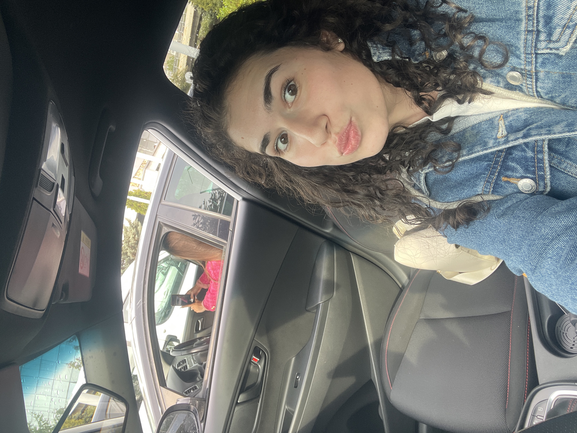 This image captures a young woman inside a car, likely taking a selfie or being photographed, with the location context provided as Sofia, Bulgaria. The photo is rotated 90 degrees counter-clockwise, showing the woman's head tilted towards the upper left.
The main subject is a young woman with dark, curly hair and light brown eyes. She is looking towards the camera with a playful "puckered lips" expression. She wears a blue denim jacket over a white top, and a small earring is visible in her right ear.
The image is taken from inside a car, showing elements of its interior. The black roof lining and a grab handle are visible at the top. The passenger door panel, with a red safety lock indicator and window controls, is on the left. Part of the black car seat, featuring red accent stitching, is seen in the lower right, with a light-colored bag resting on it.
Through the car's window on the left, an external scene is visible. Another person, wearing a bright pink top or jacket, is seen holding a smartphone, suggesting they are also taking a photo, possibly of the main subject or a selfie from outside the car. Blurred green foliage and some architectural structures are visible in the background through the windows, indicating a daytime setting. Water droplets on the window near the rearview mirror suggest recent rain or high humidity. A red warning label, partially visible with the word "WARNING," is present on the car's sun visor.