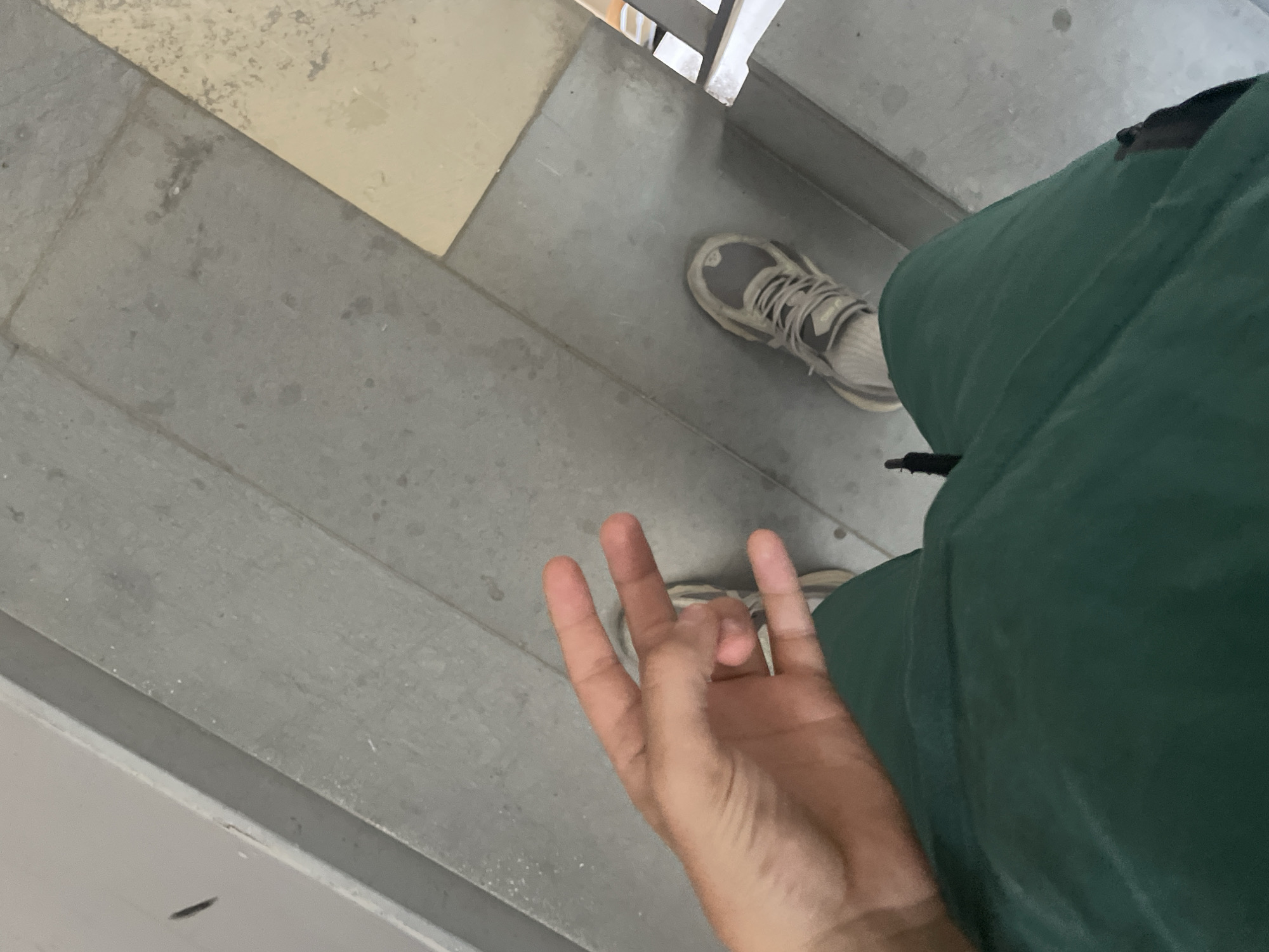 The image shows a first-person perspective of a person standing on a staircase. The subjects are the person's hand, one foot wearing a gray and white athletic shoe, and part of their green clothing. The scene is an outdoor or indoor staircase with gray tiled steps and a lighter-colored, possibly cream-colored, tile at the top left of the frame.
The person's left hand is in the foreground, with the index and middle fingers extended upwards, while the thumb and ring finger are curled inwards. The right foot, with a visible athletic shoe, is positioned further down the stairs. The green clothing appears to be pants or a jacket.
The lighting suggests it is daytime, though the weather is not discernible. There are no visible text elements in the image. The overall impression is casual and candid, with no specific event or emotion clearly conveyed. The location is suggested to be a staircase, possibly in a public building or residential area.