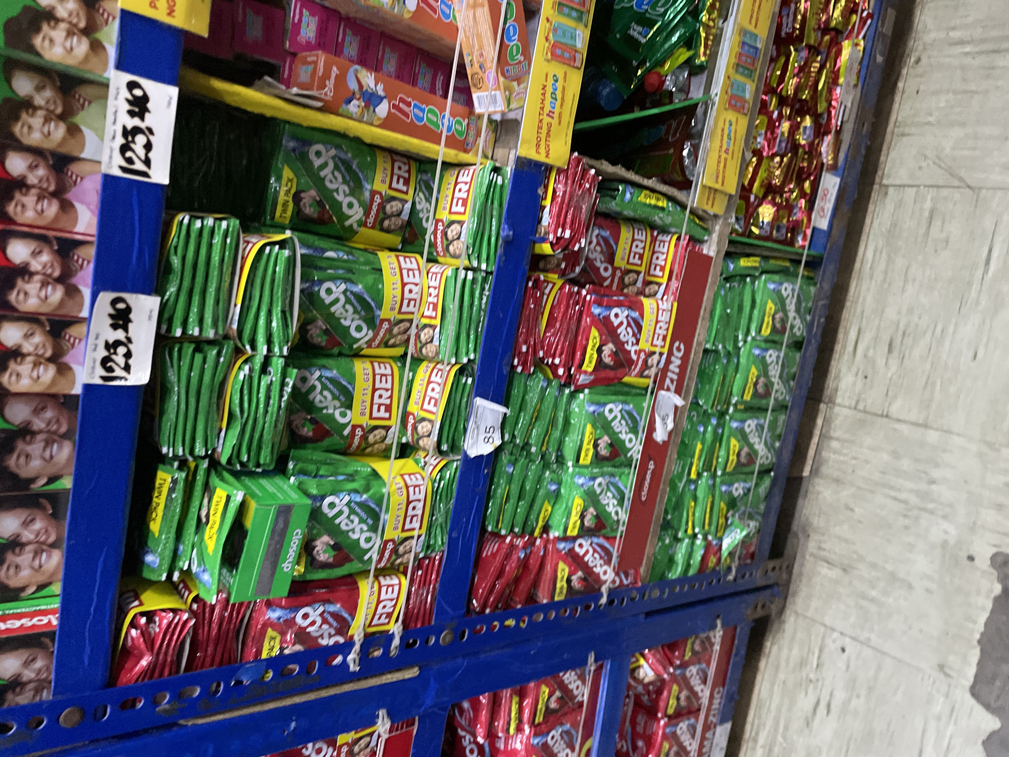 This image depicts a brightly lit interior of a retail store, likely a supermarket or grocery store, characterized by blue metal shelving units displaying a high volume of packaged goods. The linguistic elements present, such as "PROTEKTAHAN ANG NGITING hapee" (Protect the happy smile) and pricing conventions (e.g., "125.40," "85," "6.50"), indicate the location is in the Philippines. However, there are no specific markers to pinpoint the exact store name or specific address within Davao City.
The shelving unit on the left and center of the frame consists of multiple tiers. The top shelf displays large promotional posters featuring smiling young adults, with white price tags reading "125.40" affixed to the shelf edge below them. This shelf and the two shelves below it are predominantly stocked with small, flexible sachets or pouches of "Closeup" brand toothpaste. These sachets appear in two main colors: green and red. Many of the sachets, particularly the green ones, are marked with "TWIN PACK" and "BUY 1 GET 1 FREE" promotions.
On the middle shelf, a small white price tag displaying "85" is visible on the right. To the right of the main blue shelving unit, a separate display features vertically stacked colorful boxes, some adorned with cartoon characters like Donald Duck. A yellow sign above these boxes presents text that includes "PROTEKTAHAN ANG NGITING hapee" and features images of various mobile phone credit top-up denominations.
The lowest visible shelf continues to display "Closeup" toothpaste sachets, with a red shelf strip below them partially showing the word "ZINC." To the right, on a lower shelf, numerous small, shiny packages wrapped in red and gold foil are stacked. A small white price tag with "6.50" is attached near these items.
The floor beneath the shelving is covered with light-colored rectangular tiles, which exhibit visible scuff marks, dirt, and minor stains, particularly in the immediate foreground. No individuals are present in the scene, and no actions or interactions are depicted. The overall impression is that of a well-stocked and active retail environment.