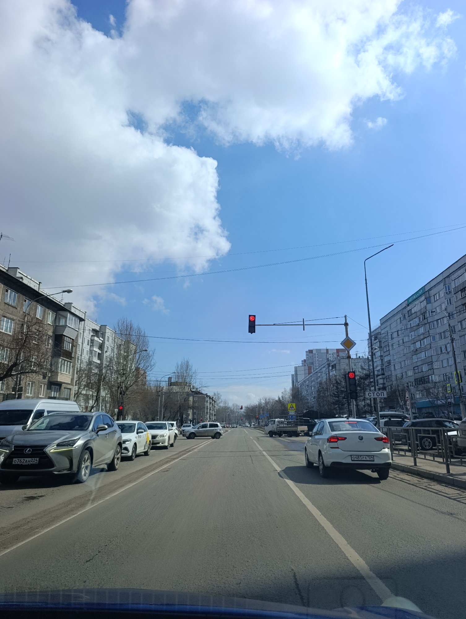 The image captures a street scene in Krasnoyarsk, Russia. The camera is positioned inside a car, looking out onto a multi-lane road. A silver Lexus SUV with license plate "O762AH224" is visible in the left lane, followed by other vehicles. A white sedan with license plate "O485PA124" is in the right lane. Traffic lights are red. Buildings line both sides of the street, with some bare trees visible. A "CTON" sign is present near the right sidewalk. The sky is blue with some clouds.