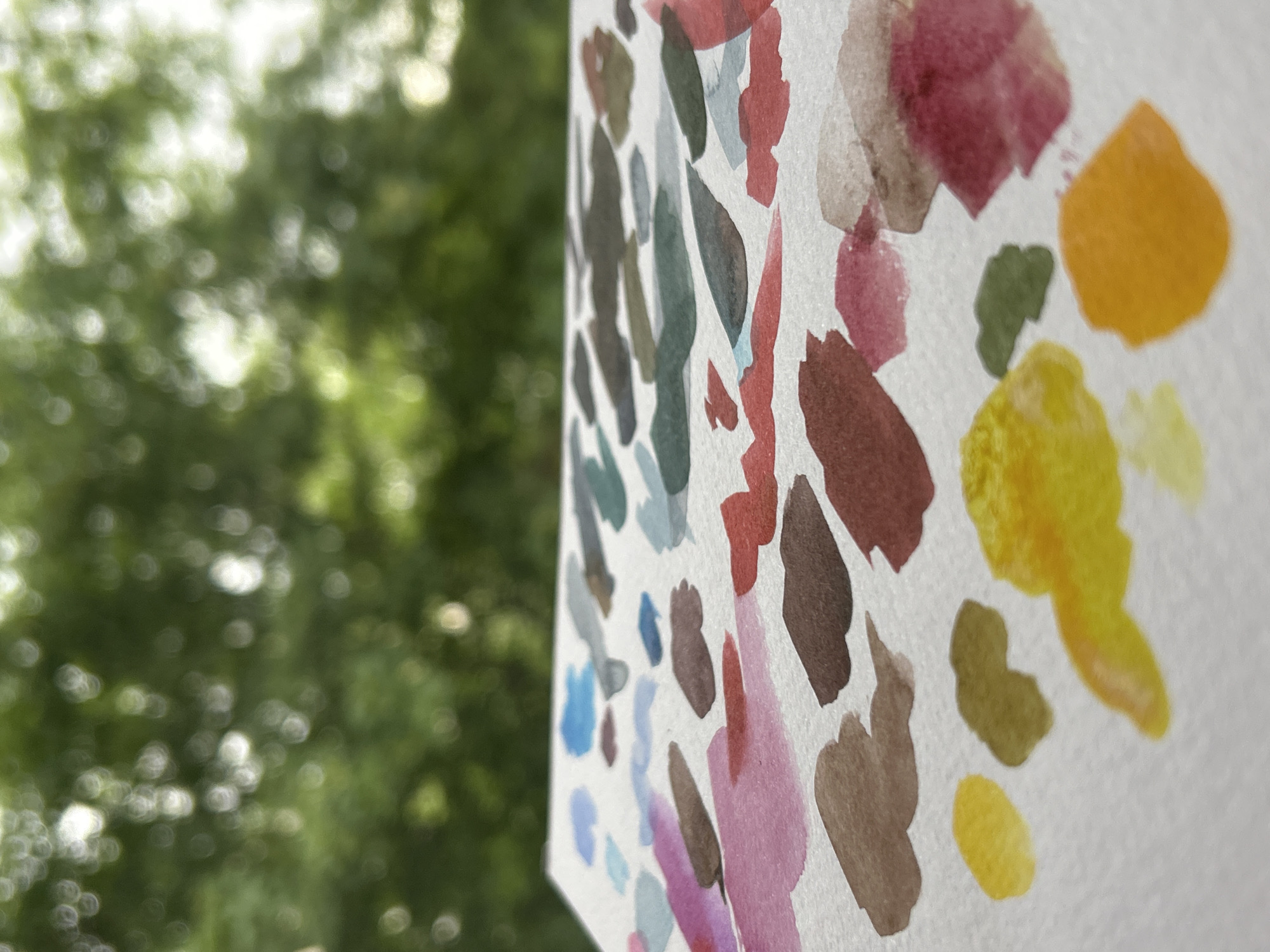 The image shows a close-up of a watercolor painting with dabs of various colors, including blues, greens, browns, pinks, reds, and yellows on a white background. The painting is held up in front of a blurred background of green foliage, likely trees, possibly outdoors. There is no indication of Sofia, Bulgaria, or any specific urban location. The focus is on the artwork and the blurred background.