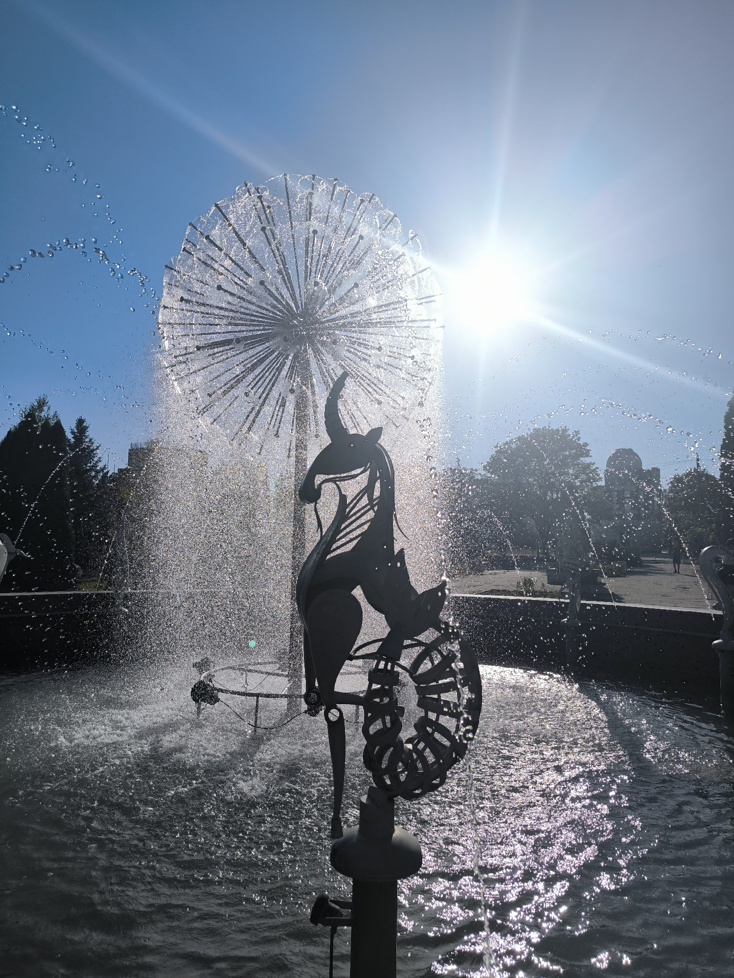 This image shows a fountain sculpture in Chișinău, Moldova. The sculpture appears to be a metal abstract depicting a creature, possibly a dragon or griffin, with large wings or fins. It is positioned in front of a dandelion-shaped water feature that sprays water in a spherical pattern. The sun is bright and high in the sky, creating lens flare and illuminating the water droplets, making them sparkle. In the background, there are trees, a path, and buildings, suggesting a park or public square setting. The water from the fountain is creating a mist and reflecting the sunlight. There are no people clearly visible in the immediate foreground, though a distant figure can be seen walking on the path in the background. The overall scene is one of a bright, sunny day at a decorative fountain.