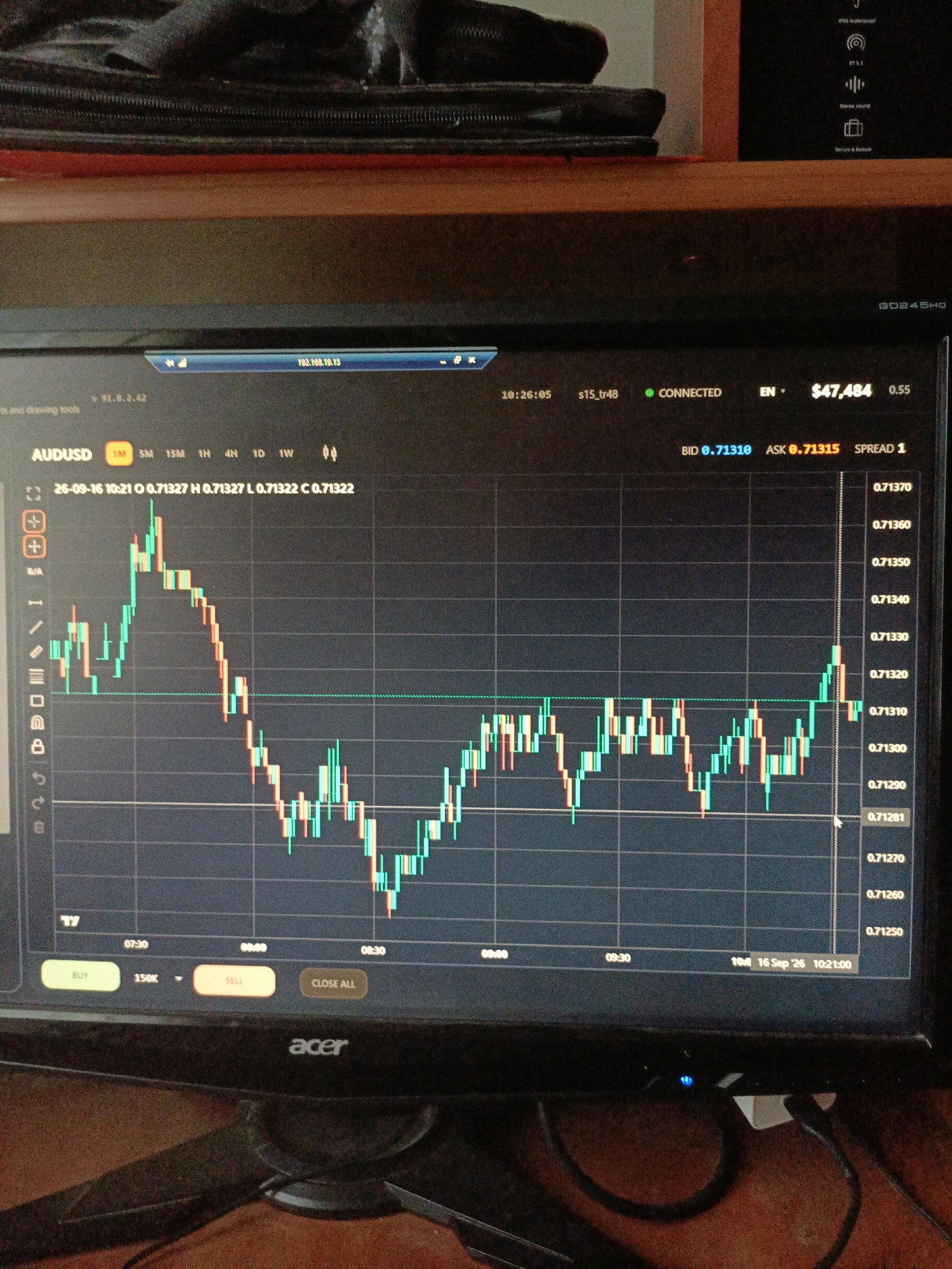 The image shows a close-up of a computer monitor displaying a financial trading chart. The chart shows candlestick patterns representing price movements of AUDUSD. The monitor is placed on a wooden desk, with some cables visible at the bottom. In the background, a black zippered bag is placed on a red surface. The monitor displays a timestamp of 10:26:05 and the date 26-09-16. It also shows the current Bid of 0.71310 and Ask of 0.71315. The image appears to be taken indoors.