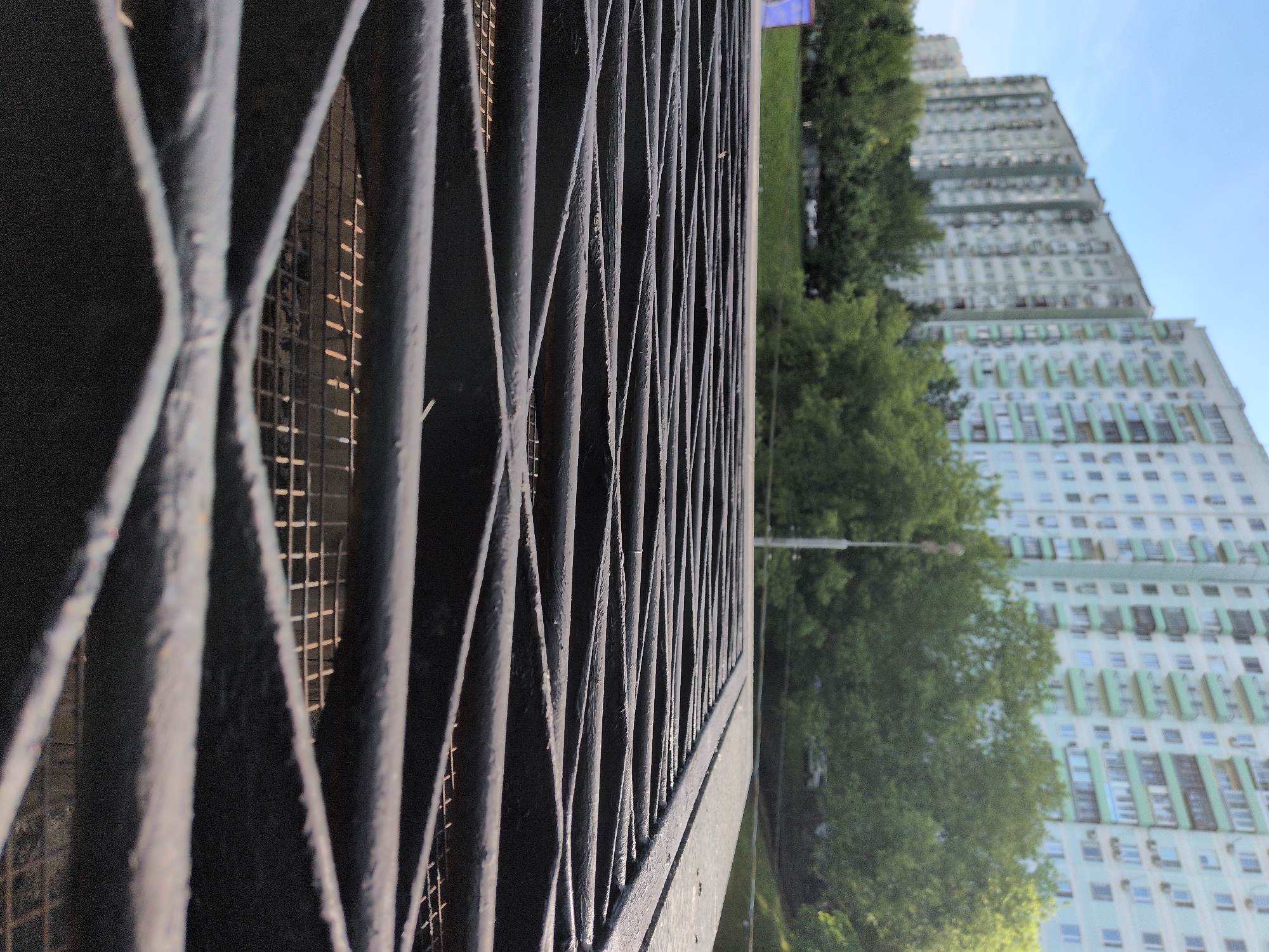 The image shows a metal fence with a grid pattern, through which a high-rise apartment building and trees are visible. The building has a light color scheme with many windows. The scene suggests an urban environment. The location is identified as Moscow, Russia.