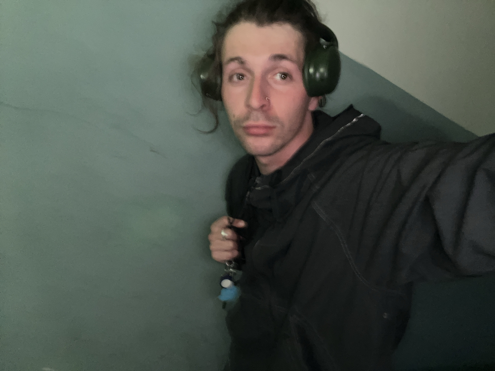 A selfie depicting a man with brown hair pulled back, wearing green earmuffs and a black hooded jacket. He has a nose ring and is holding keys with a small blue keychain. The background is a pale green wall with a sloped ceiling, creating a cramped or enclosed space. The location is not discernible from the image.