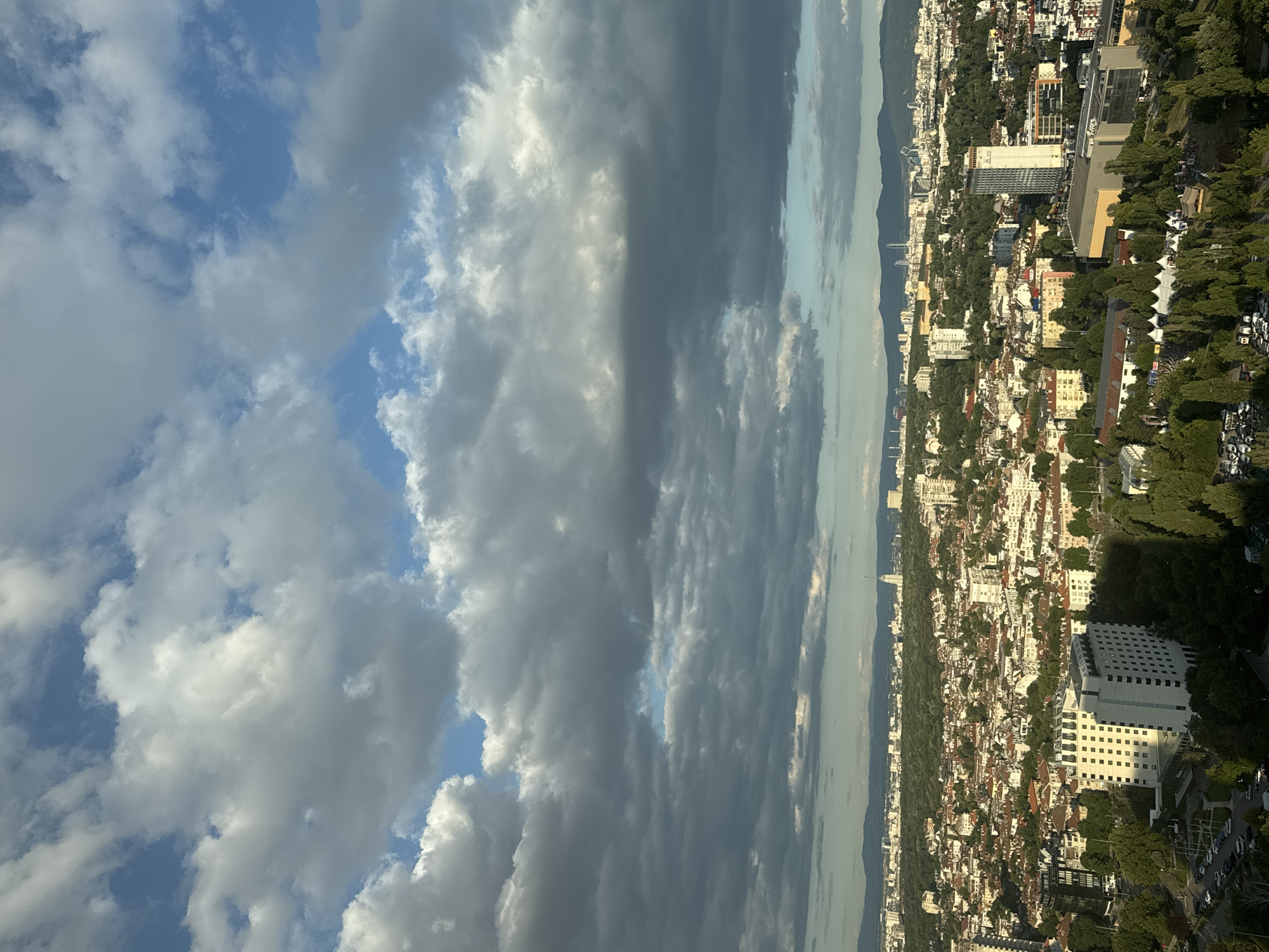 This is an aerial view of the city of Sofia, Bulgaria, on a partly cloudy day. In the foreground, there are several modern buildings surrounded by lush green trees. One prominent white building has a rectangular shape with many dark square windows. To its left, a darker building with a similar window pattern is visible. Further into the scene, the dense urban landscape of Sofia unfolds, characterized by a mix of residential buildings with red-tiled roofs and taller, modern structures. In the distance, a mountain range forms the horizon. The sky is filled with dramatic clouds, some dark and heavy, others lighter and scattered, with patches of blue sky peeking through. The lighting suggests it is either early morning or late afternoon, casting shadows across the cityscape. There are no people or discernible actions visible in the image.