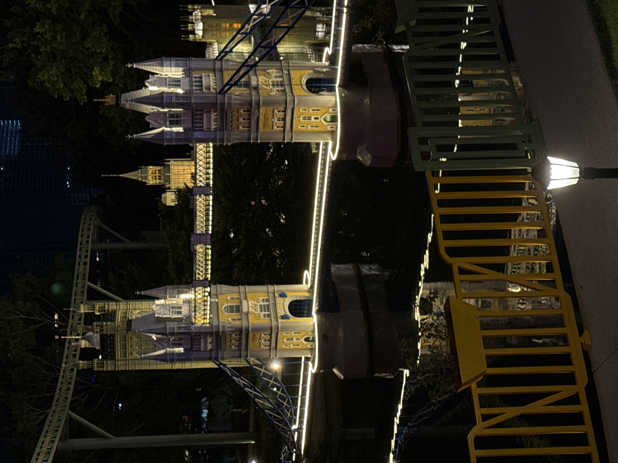 The image depicts a nighttime scene of a miniature replica of the Tower Bridge in London, situated within a park in Nanshan District, China. The replica is illuminated, highlighting its architectural details, including the twin towers and the bridge's suspension cables. The bridge spans a body of water, reflecting the lights. In the background, behind the replica, trees are visible, and further back, the lights of a modern cityscape suggest the presence of tall buildings. In the foreground, a pathway is bordered by a colorful fence made of yellow and green metal bars, with a modern lamppost casting light onto the path. The scene appears to be a tourist attraction or a themed park. There are no people visible in the image.