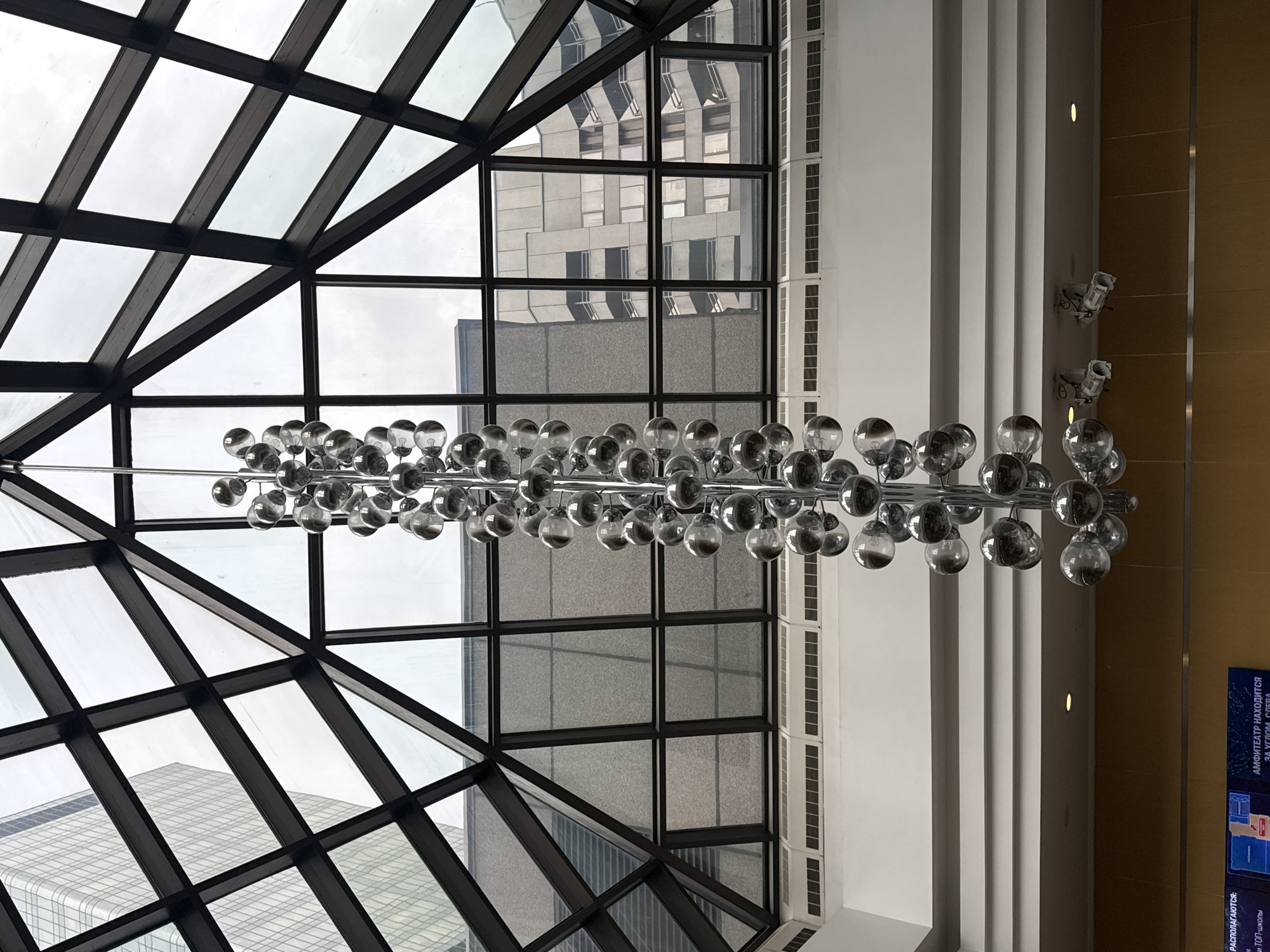 The image shows the interior of a building with a glass-paneled, angular skylight. Outside the skylight, other buildings are visible, suggesting a city environment. A modern chandelier with numerous spherical lights hangs suspended in the interior space. The architectural style is contemporary with white pillars and wood paneling visible on the walls. A sign in Russian is visible on the right. Based on the architectural style and the presence of a Russian-language sign, the location is Moscow, Russia.