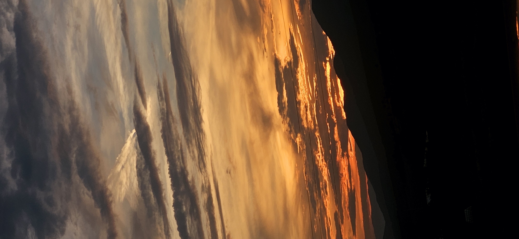 A vertical, outdoor photograph captures a dramatic sunset over a mountainous landscape in Momin Prohod, Bulgaria. The sky is filled with a vivid gradient of colors, transitioning from deep oranges and reds near the horizon to softer yellows and grays higher up. Wispy, horizontal clouds are illuminated by the setting sun, creating a sense of depth and movement. Silhouetted against the colorful sky are the dark, undulating shapes of mountains. The foreground is obscured by darkness, suggesting a nighttime or twilight setting. No people or specific structures are clearly discernible in the image. The overall impression is one of natural beauty and a serene, twilight atmosphere.