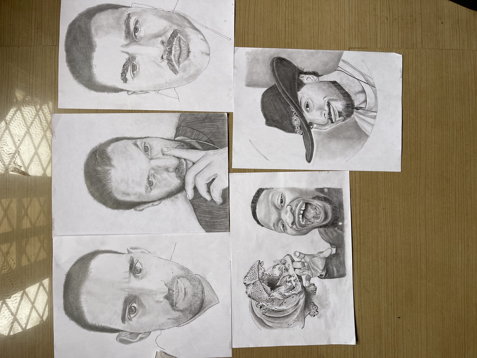 The image shows five graphite pencil drawings on white paper, arranged on a corrugated cardboard surface. The drawings appear to be portraits of men in various poses. One subject is wearing a cowboy hat. Another subject is pointing a finger toward their face. Another subject is drawn with their mouth wide open, revealing teeth and tongue. A close-up of hands holding a detailed rendering of what may be a sea creature is included in the lower-right. No aspects of the image directly indicate a specific location in Jos, Nigeria.