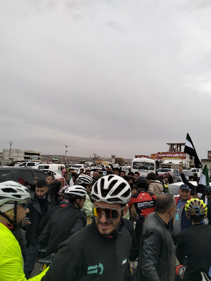 A crowd of cyclists, mostly wearing helmets and cycling gear, gathers in a parking lot. Many wear casual attire. Some hold black and white flags. A white van and multiple cars are parked behind the crowd. Buildings are visible in the background under an overcast sky. The cyclists are likely at the starting point of a race.