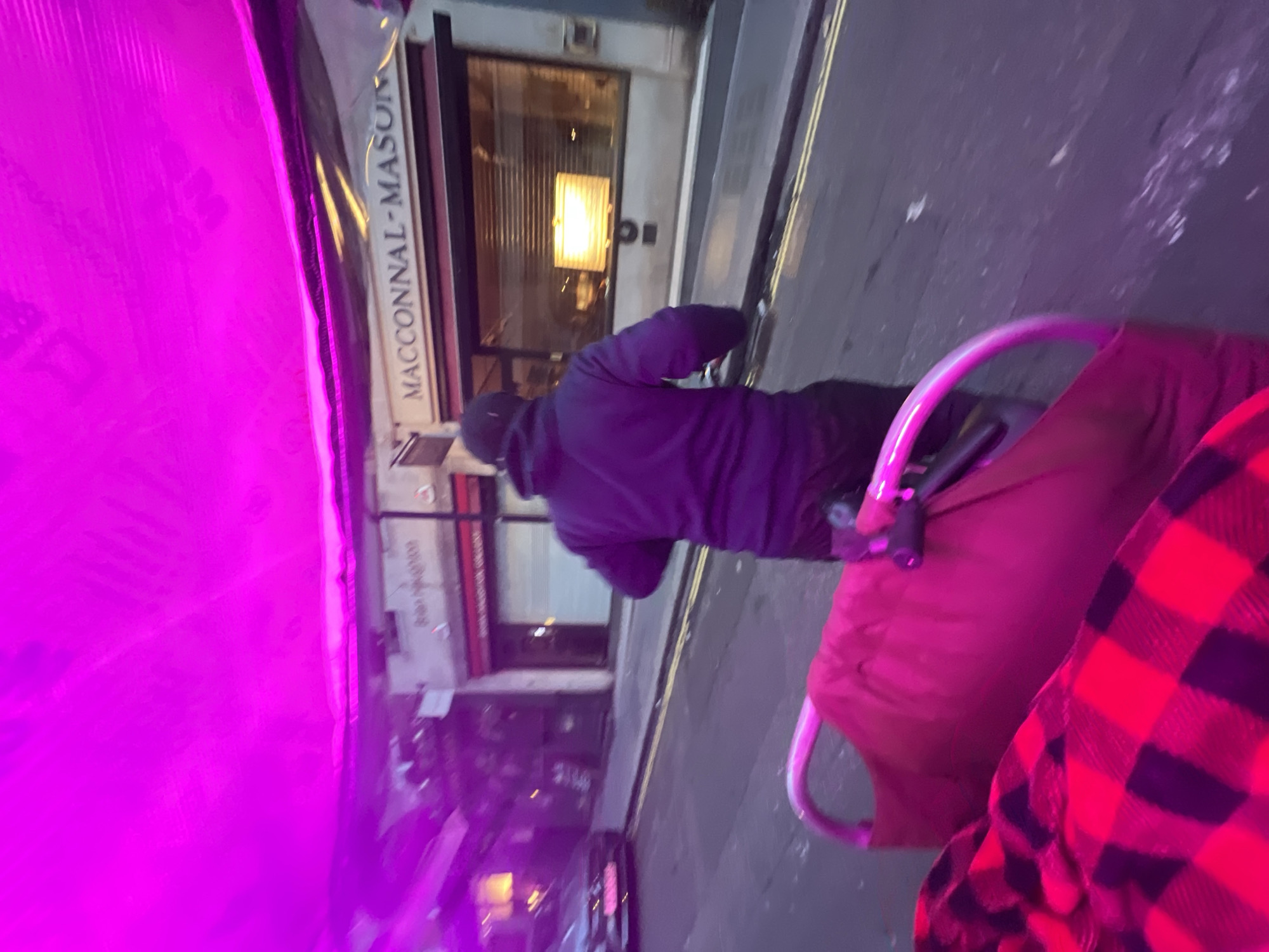 A street in the city of London, United Kingdom, shows a man wearing a hooded jacket walking down the sidewalk past a building with the name "MACCONNAL-MASON" visible on its facade. He appears to be pushing a baby stroller, partially obscured by what may be a red and black plaid blanket or garment. The scene is shot from an elevated perspective with an artificial lighting source causing a strong pink hue to permeate parts of the image.