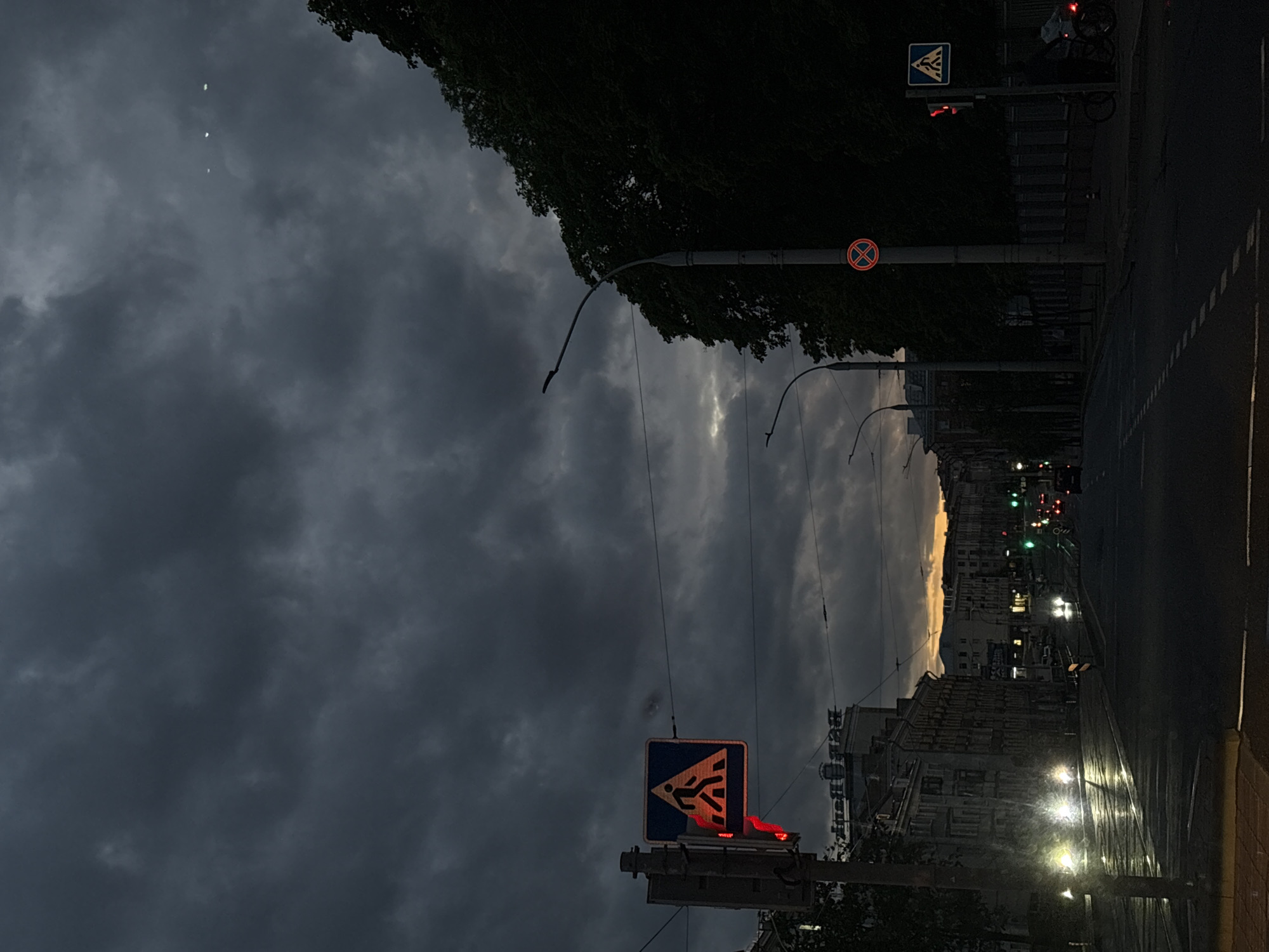 The image depicts a city street at dusk, likely Minsk, Belarus, given the architectural style of the buildings. Overcast skies dominate the scene. Traffic lights are visible, some showing red. Streetlights illuminate the wet street. Pedestrian crossing signs are present. Buildings line the street, with lights on in some windows. Cars with headlights on move along the street. There are some trees that could be used for shade and some wires running above the street.
