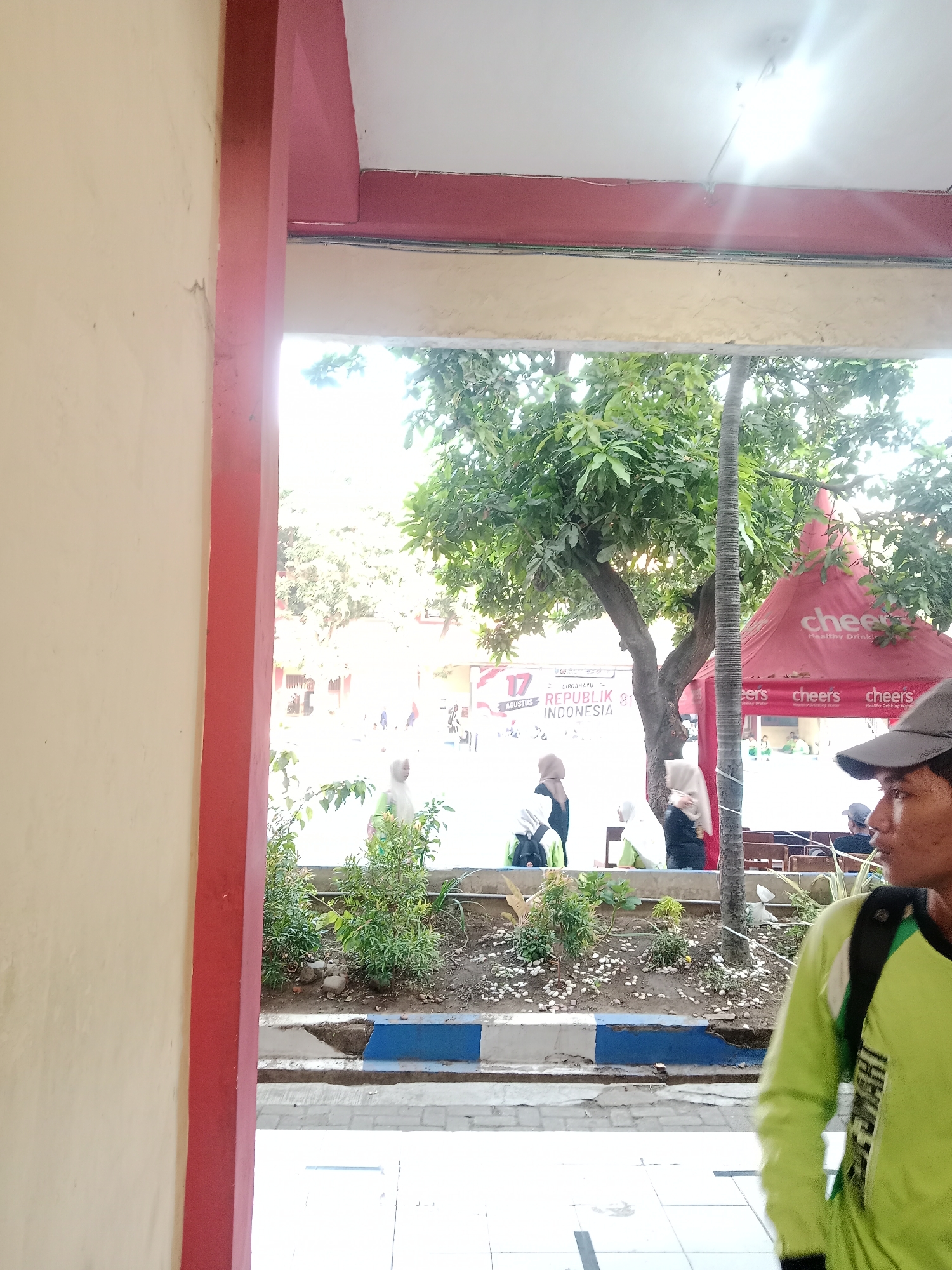 The image captures an outdoor scene, possibly a street or public area, in Winong, Indonesia. In the foreground, on the right side, a young man wearing a bright green long-sleeved shirt and a dark backpack is partially visible, his face turned to the right. To the left, a wall painted a light cream color with a vertical red beam is prominent. Beyond the wall, a group of people, some wearing hijabs, are seen in the midground. They appear to be gathered near a red tent with "cheers" branding. Behind them, a tree with lush green foliage is visible. The ground in the midground consists of soil with some small white pebbles and green plants, bordered by a blue and white stripe. The background shows a building and a banner with text that includes "17 AGUSTUS REPUBLIK INDONESIA". The lighting suggests it is daytime, with sunlight filtering through the trees and a bright light source at the top of the image, possibly an overhead lamp.