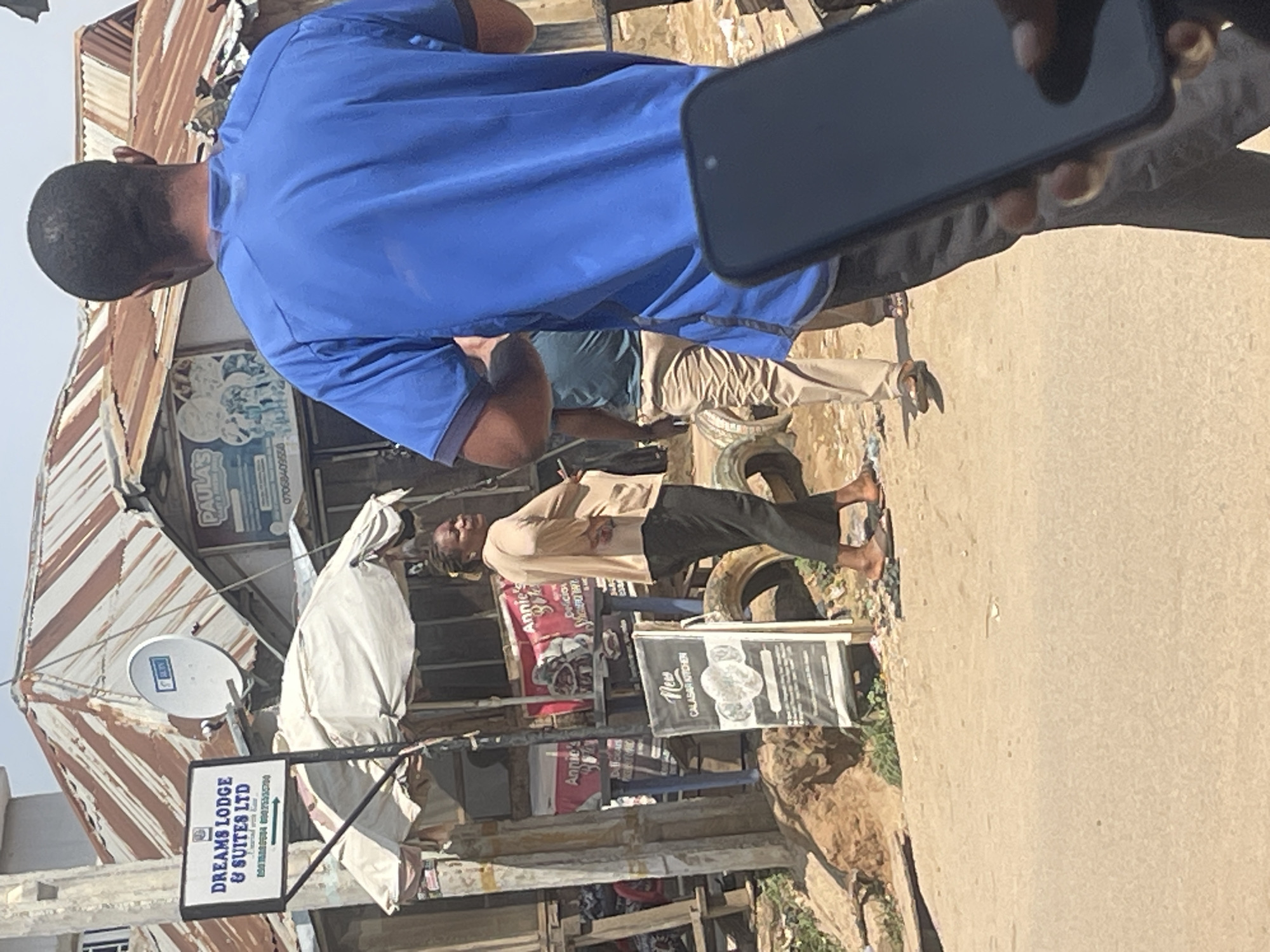 This image captures a street scene in Abuja, Nigeria, likely during the day given the bright lighting. The focus is on a group of people in an outdoor setting, possibly a market or a commercial area. The foreground is dominated by a person wearing a bright blue shirt, with their back to the camera. To the left, a structure with a corrugated metal roof, showing signs of rust, suggests a somewhat informal or weathered commercial establishment. A satellite dish is mounted on this structure.
Further into the scene, another person is visible in a light-colored shirt and dark pants, standing with their arms crossed and looking upwards. Behind them, a large tire is propped up. Several signs are visible, including one for "DREAMS LODGE & SUITES LTD" and another that appears to advertise "Annie's" and "New CALASAR KITCHEN". The presence of these signs, along with the general atmosphere of the scene, points towards a commercial district or a busy thoroughfare where small businesses operate. The ground appears to be paved. A hand holding a dark-colored smartphone is partially visible in the top right corner, suggesting someone might be taking a photograph or recording the scene. The overall impression is one of everyday life unfolding in an urban environment.