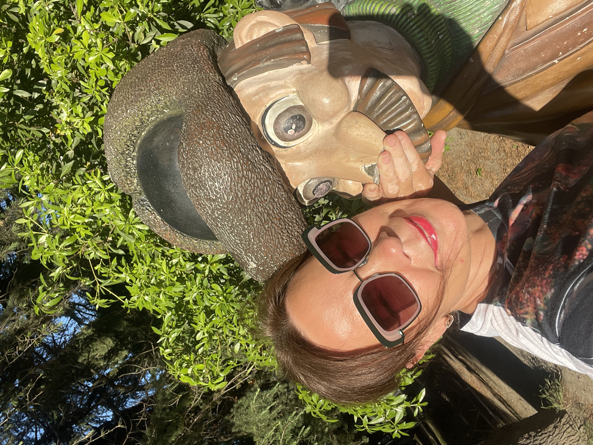 A woman with short brown hair, wearing oversized sunglasses and a black t-shirt with a colorful abstract print, poses for a selfie next to a whimsical statue of a man wearing a large brown hat and a green scarf. The statue has a cartoony appearance with large eyes and a wide smile, and one of its hands is covering its mouth. The woman's left hand is also near the statue's face. They are in an outdoor setting with lush green foliage and trees behind them, suggesting a park or garden. Sunlight casts shadows on the ground and on the statue. The scene appears to be in a city park in Sochi, Russia, possibly in the Arboretum Botanical Garden, known for its diverse plant life and decorative elements.