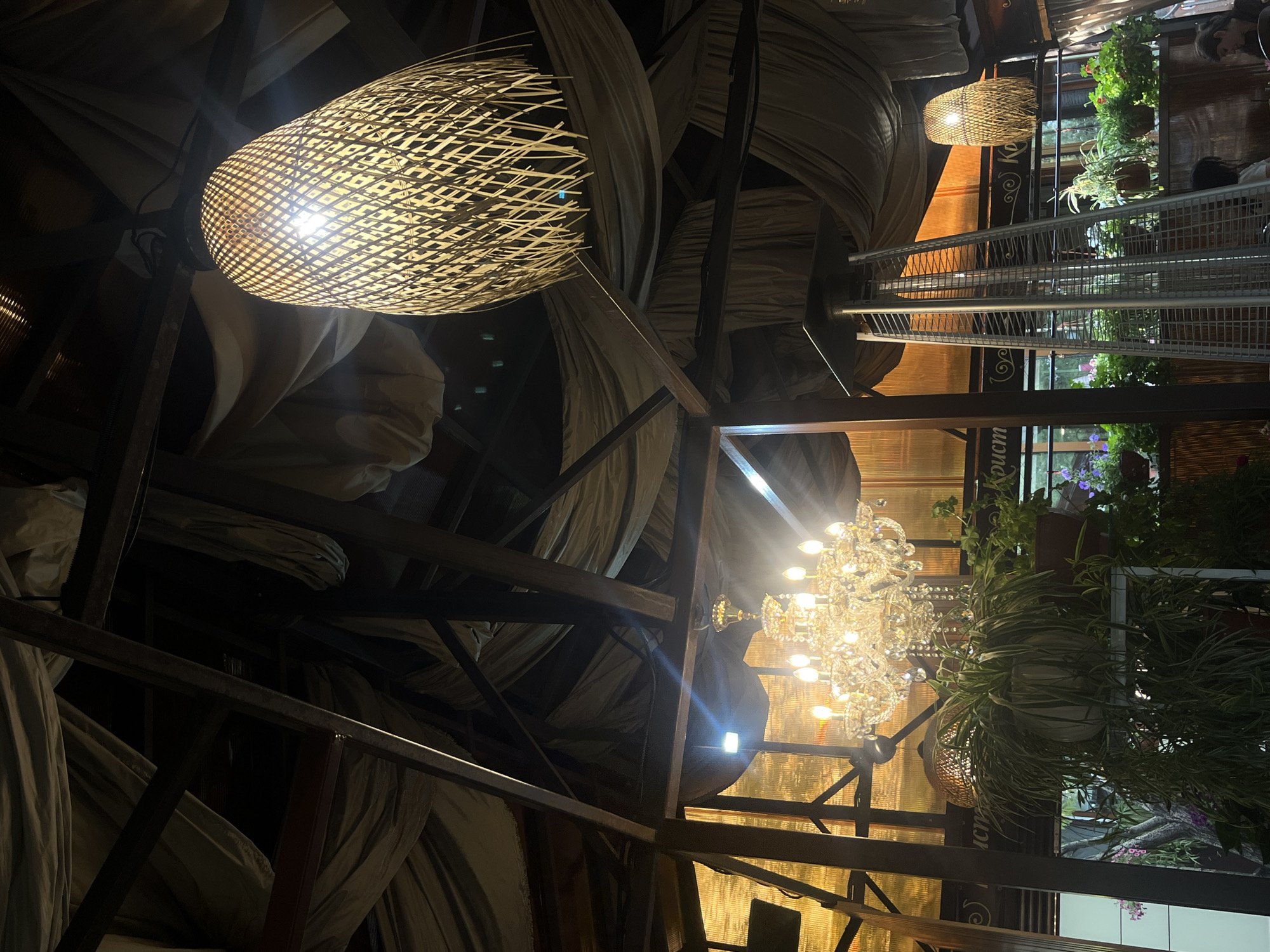 The image depicts the interior of a dimly lit establishment, possibly a restaurant or cafe. Dominating the foreground is a large, woven basket-style lampshade with a bright bulb inside, casting a warm glow. Behind it, a dramatic chandelier with multiple crystal elements and lit bulbs hangs, reflecting light. A metal structure, possibly shelving or part of the building's framework, is visible, interspersed with flowing, draped fabric in shades of grey and beige.
In the background, there are glimpses of greenery, suggesting the presence of potted plants or a garden area. A metal railing or shelf unit is also visible, holding more plants. Faint outlines of people can be seen in the distance, indicating that the establishment is occupied. The overall ambiance is one of subdued lighting and an eclectic decor, with a mix of natural materials and ornate fixtures. The time of day appears to be evening or night, given the artificial lighting. There is no indication of weather conditions.
Visible text in the image includes "Константинополь" written on a decorative element behind the chandelier. The location is identified as Novy Urengoy, Russia.