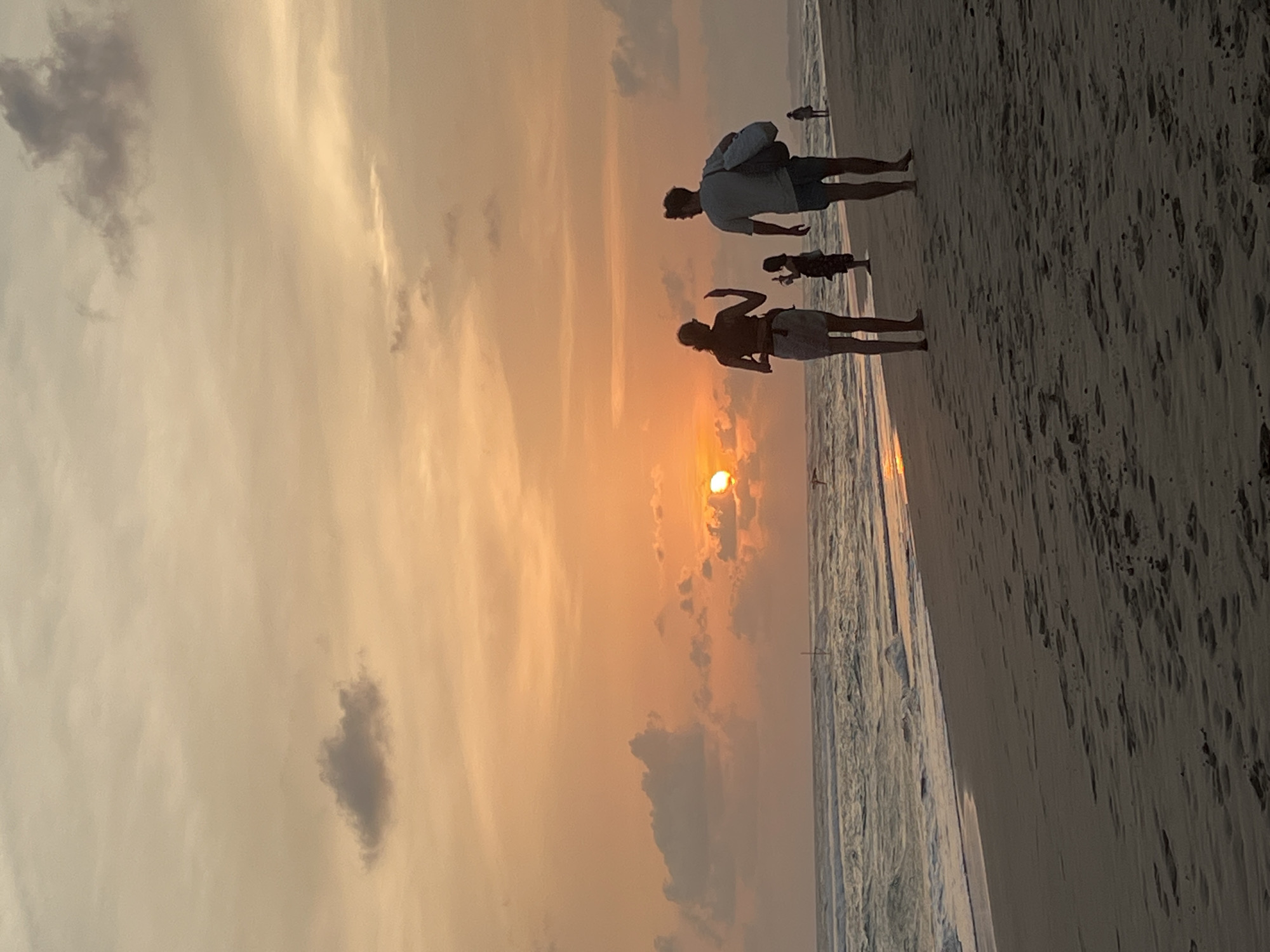 The image captures a sunset scene at Kathaluwa, Sri Lanka beach. Silhouettes of three people are visible. A man in a light-colored shirt is carrying a bag. Next to him, a child-sized figure stands closer to the shore. A woman with a dark upper garment is located closest to the setting sun. The sun is partially obscured by clouds, creating a warm, golden glow in the sky. The beach stretches into the foreground. The ocean reflects the sunset colors.