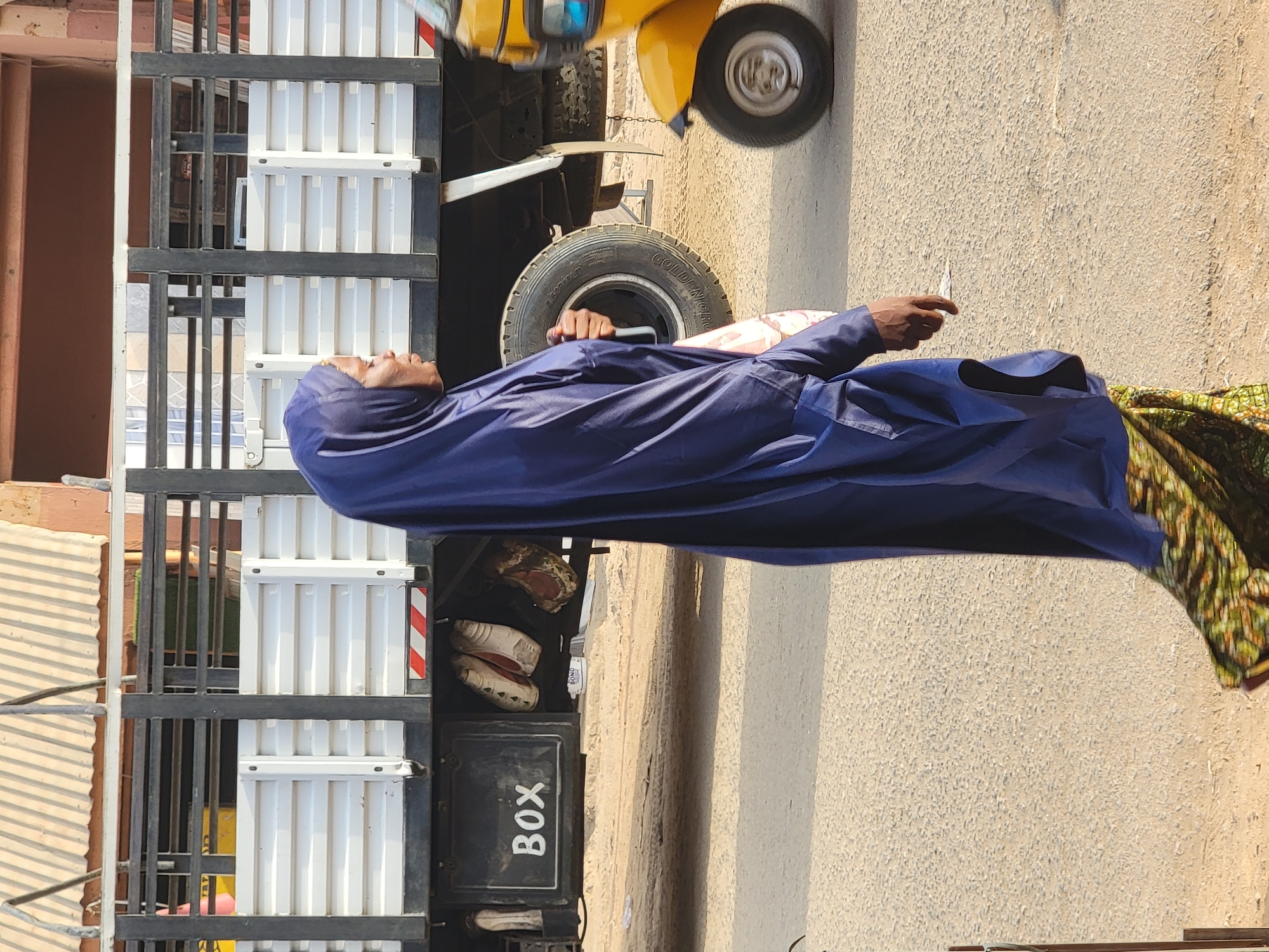 A woman in a blue garment and head covering stands on an asphalt road in Maiburiji, Nigeria, next to a commercial truck with white corrugated panels and black metal frame. A yellow three-wheeled vehicle is parked nearby. The back of the truck includes a storage box labeled "BOX." Items such as shoes and a white bottle with labeling are visible in a space beside the box. The woman is holding a small white object in her right hand. The road surface is light tan and shows signs of wear.